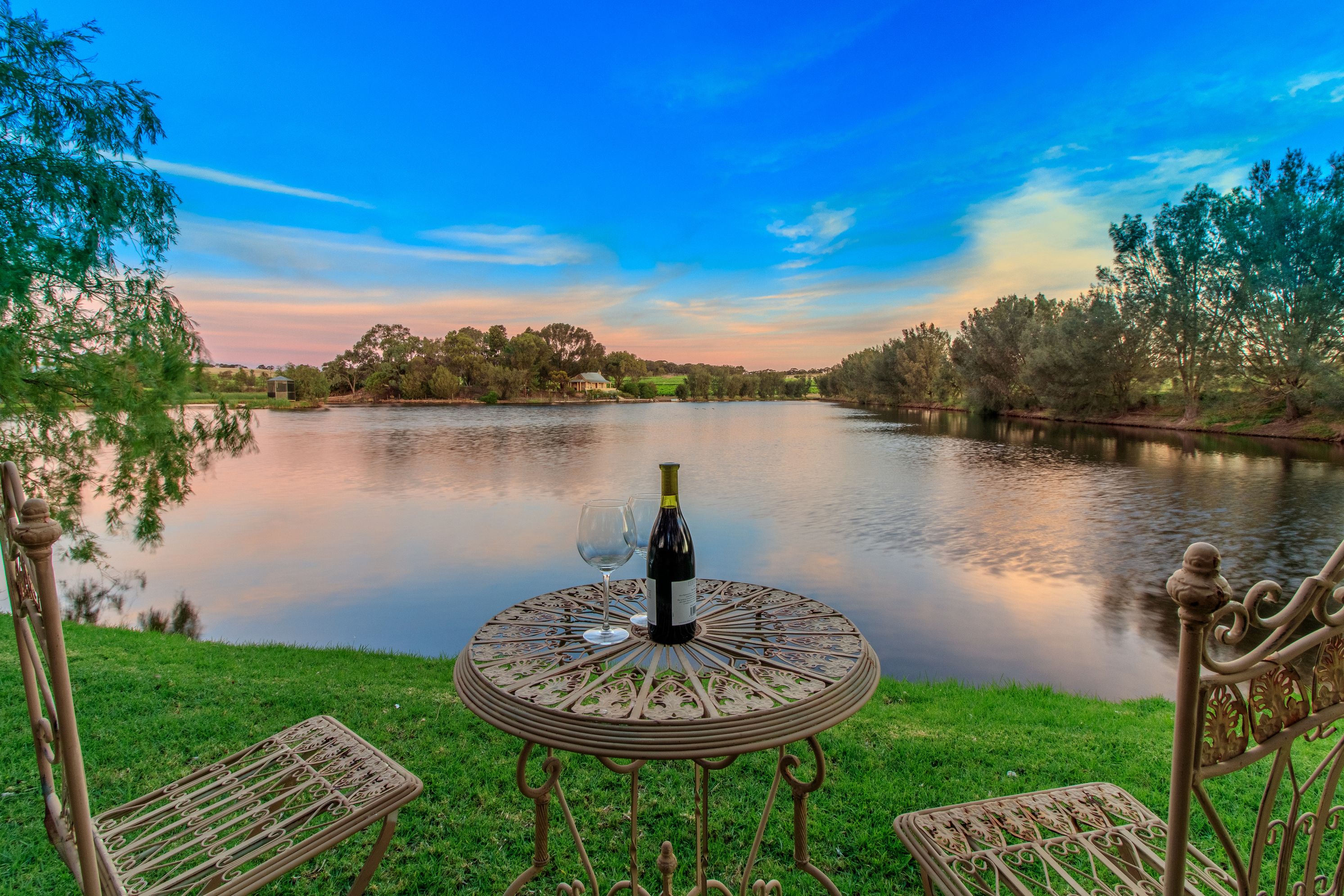 Mesa redonda de metal com uma garrafa de vinho e um copo, colocada numa área gramada perto de um lago calmo com árvores ao fundo.