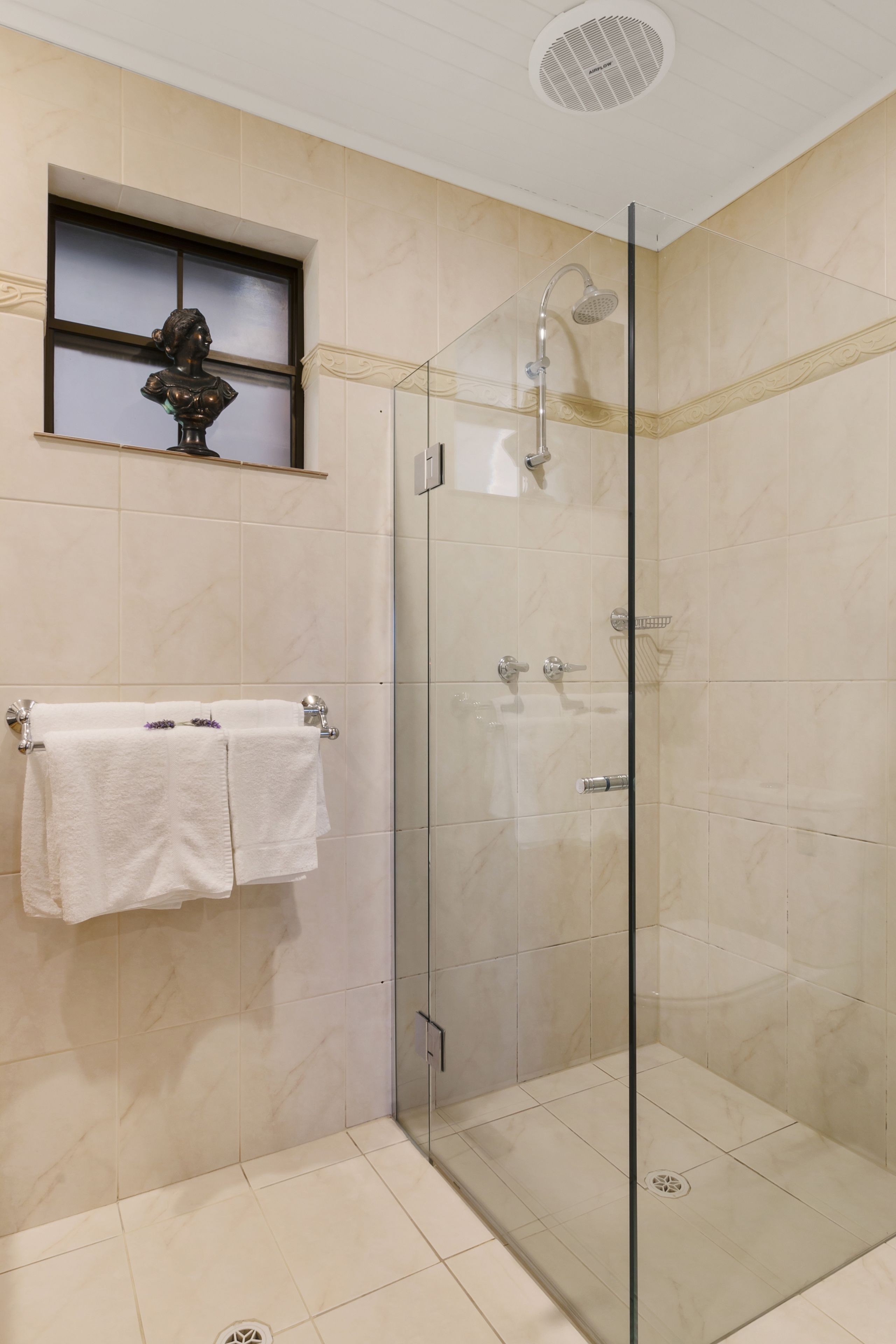Une douche fermée par des parois en verre, dotée d’un pommeau de douche à main, d’un porte-serviettes avec des serviettes blanches et d’un buste décoratif accroché au mur.