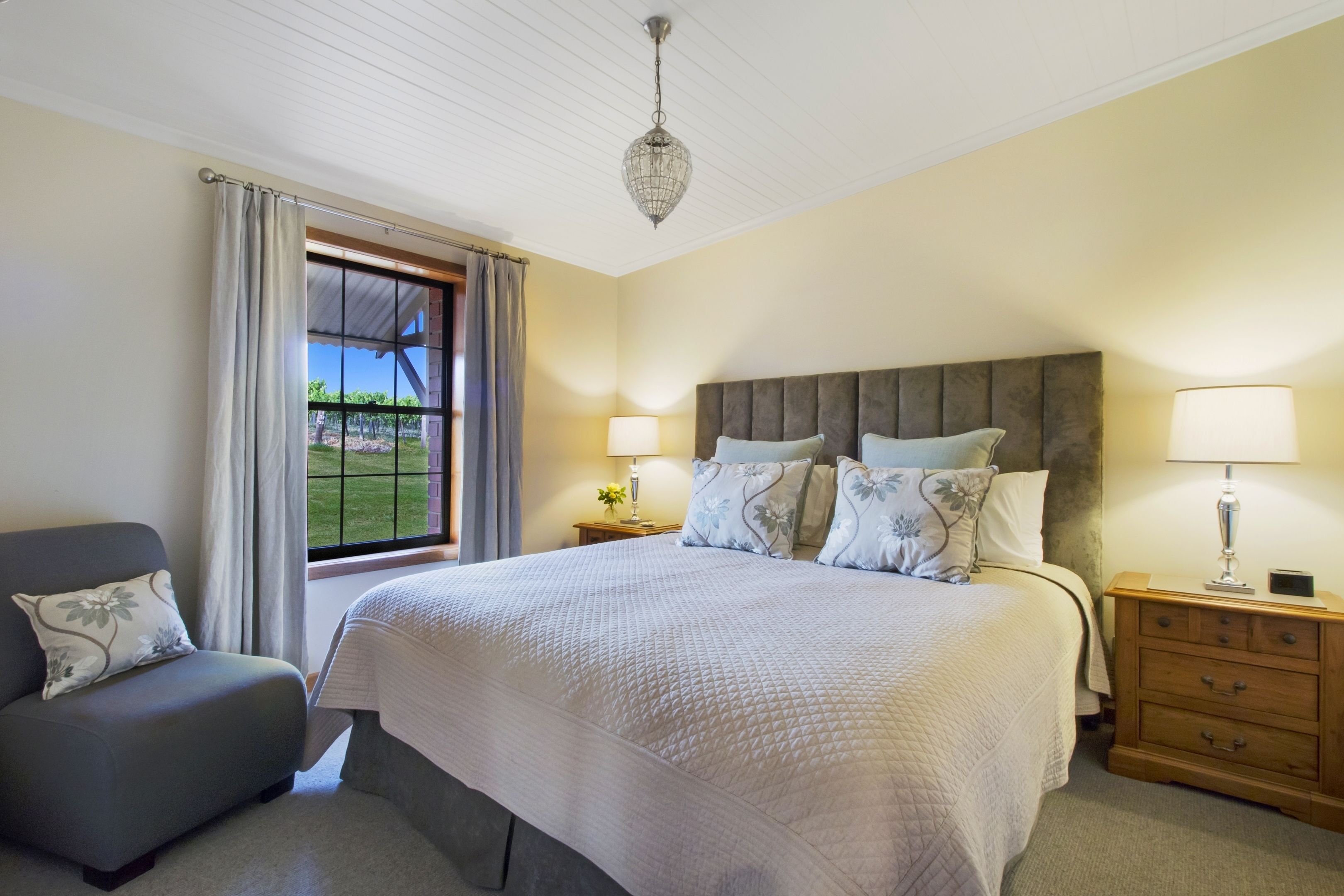 A bedroom with a large bed, a nightstand, a lamp, a chair, and a window with curtains.