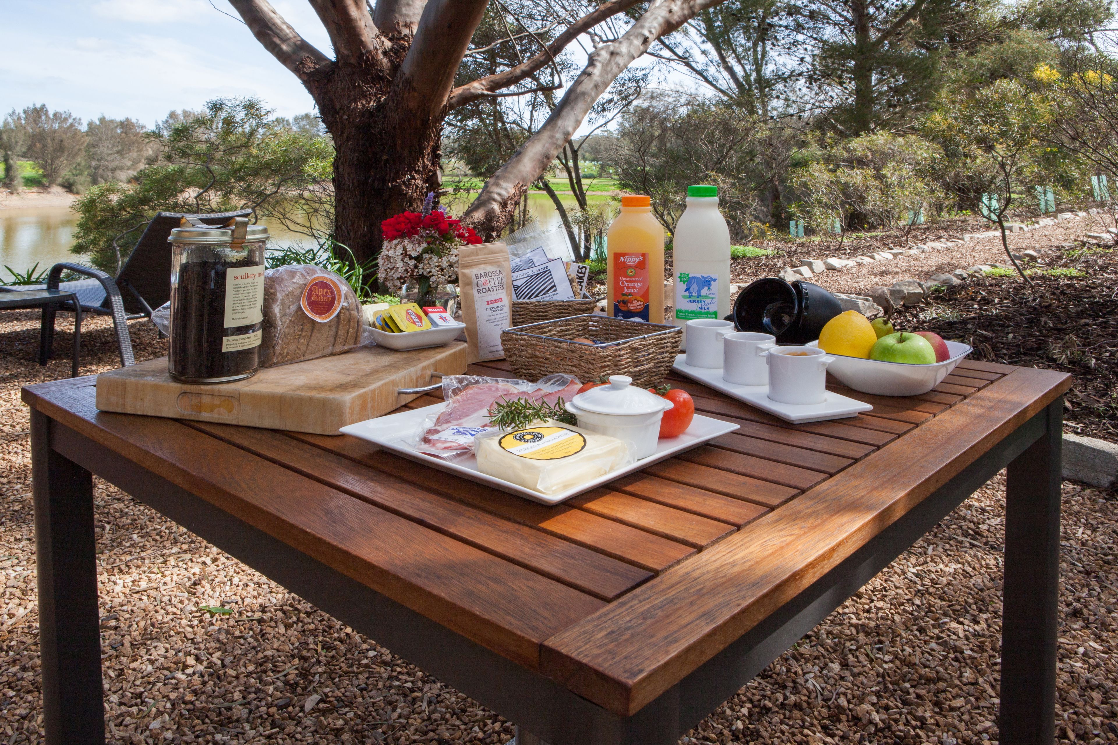 A wooden table set with breakfast items including bread, cheese, eggs, and fruits, with a view of trees and a body of water in the background.
