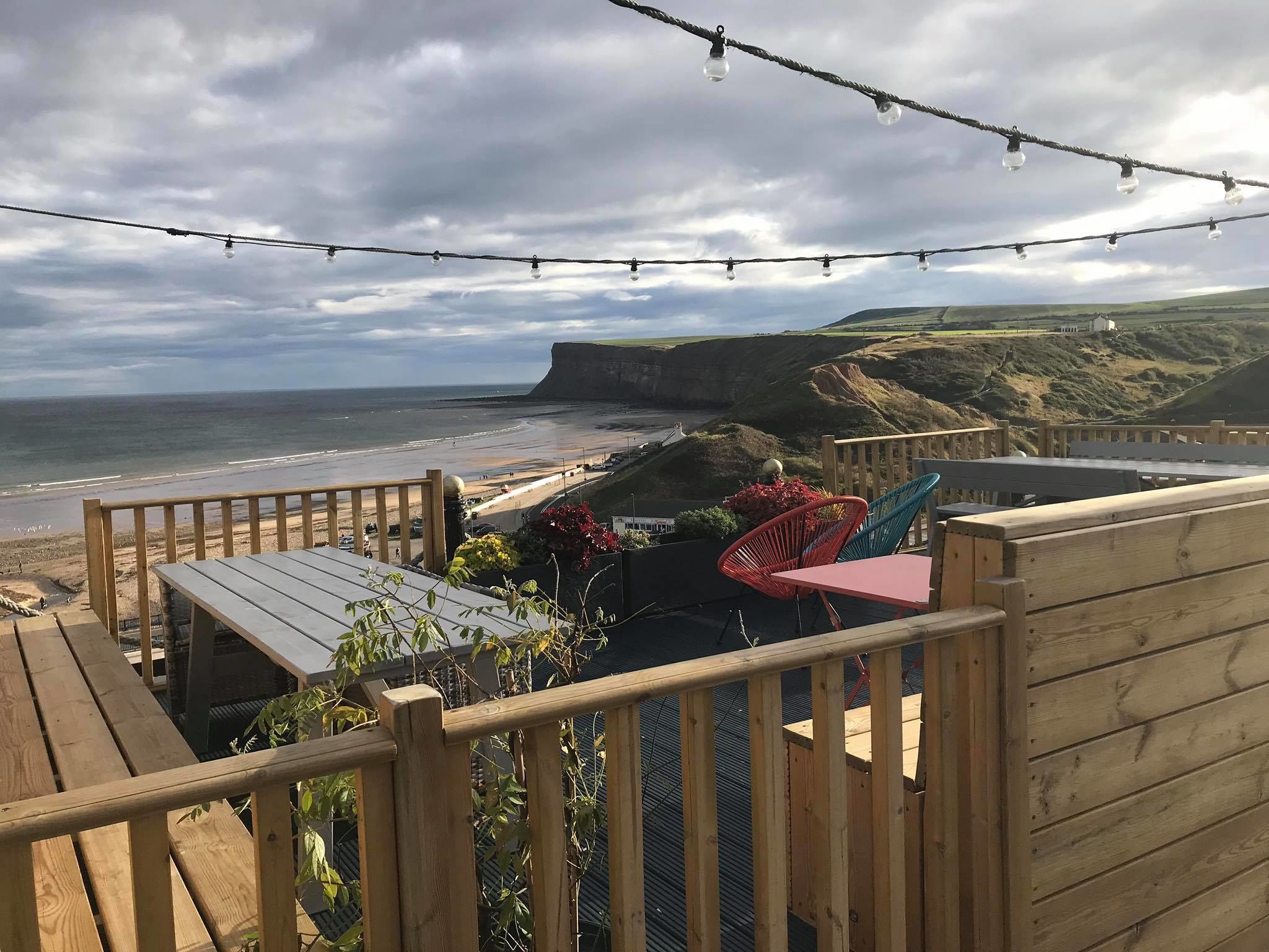 Une terrasse en bois avec une table et des chaises, donnant sur une plage et des falaises.