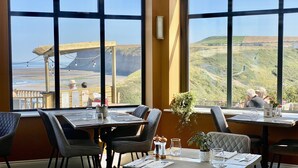 A restaurant with large windows offering views of a coastal landscape, including a beach and hills. Tables are set with glasses and cutlery, and there are potted plants on the windowsill. - The Spa Hotel (Saltburn-by-the-Sea)