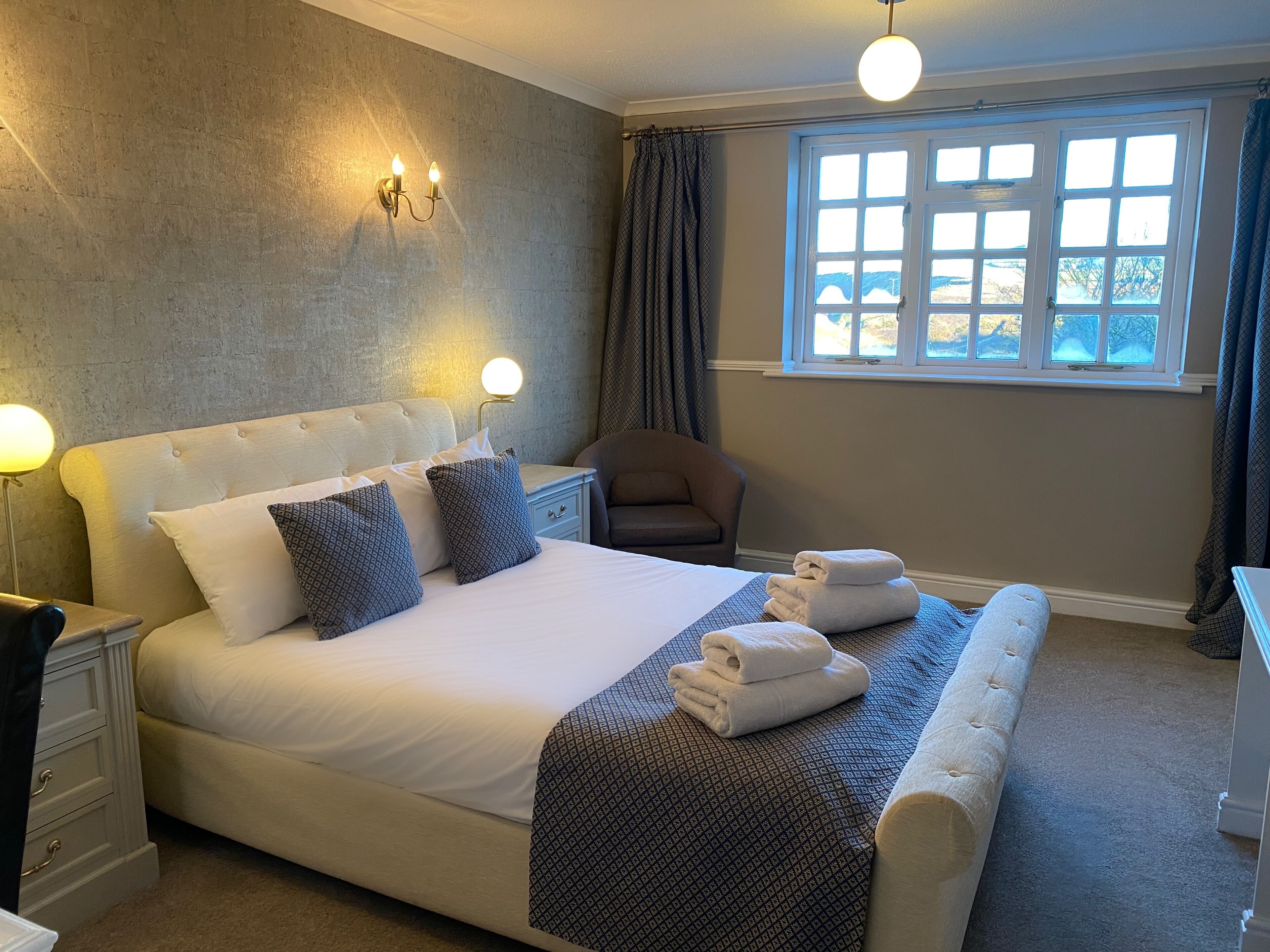 A neatly arranged bedroom with a large bed, bedside lamps, a chair, and a window with curtains.