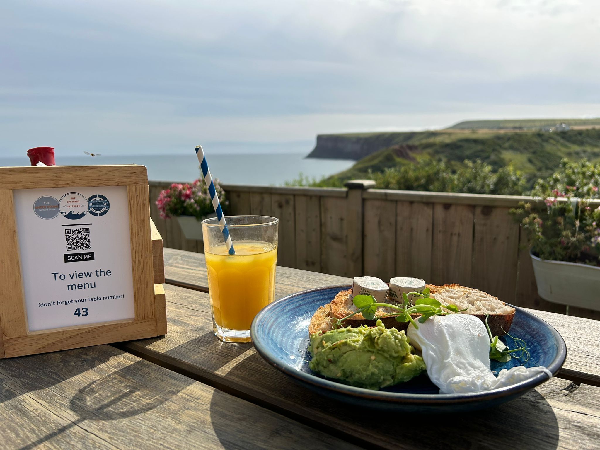 A table with a QR code menu, a glass of orange juice, and a plate with avocado and poached egg on a wooden deck overlooking the sea.