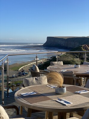 A seaside terrace with round tables and wicker chairs, overlooking a beach and cliffs. - The Spa Hotel (Saltburn-by-the-Sea)