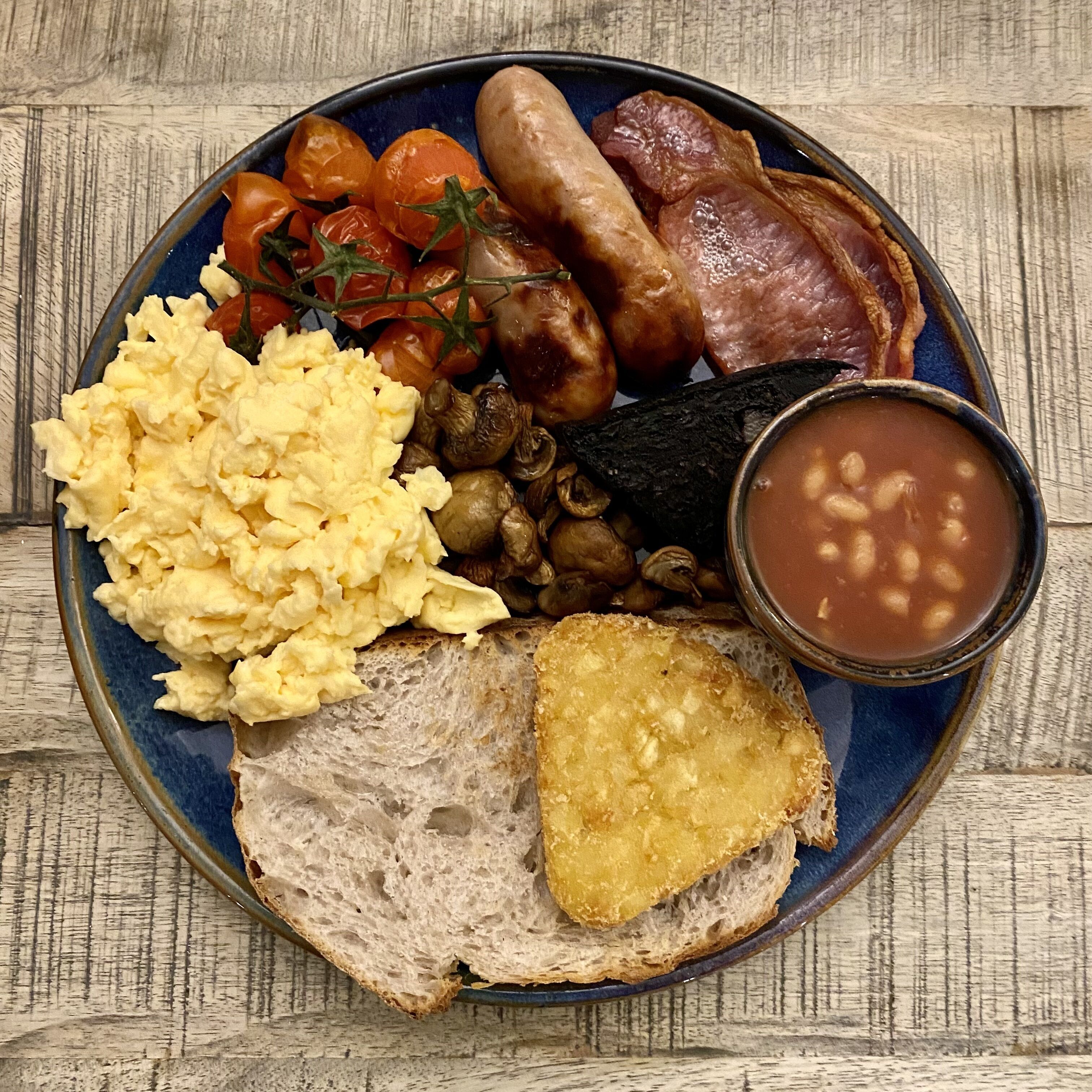 Un plato con huevos revueltos, salchichas, tocino, tomates asados, champiñones, frijoles, tostadas y un tazón de sopa.