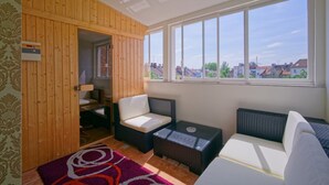 A sunlit room with a wooden wall, a sofa, a small table, and a view of buildings through large windows. - Penzion Janka (Brno)