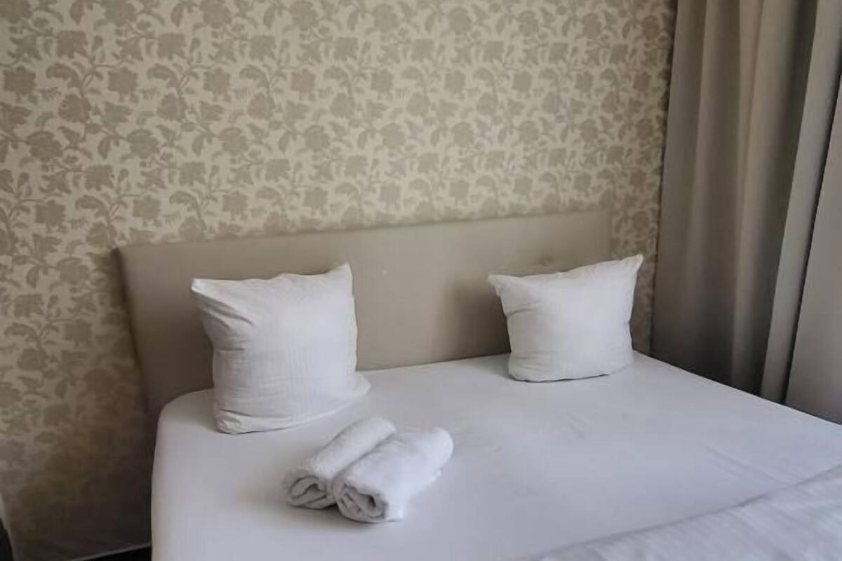 A neatly made bed with white linens and pillows, a floral-patterned wallpaper, and a window with curtains.