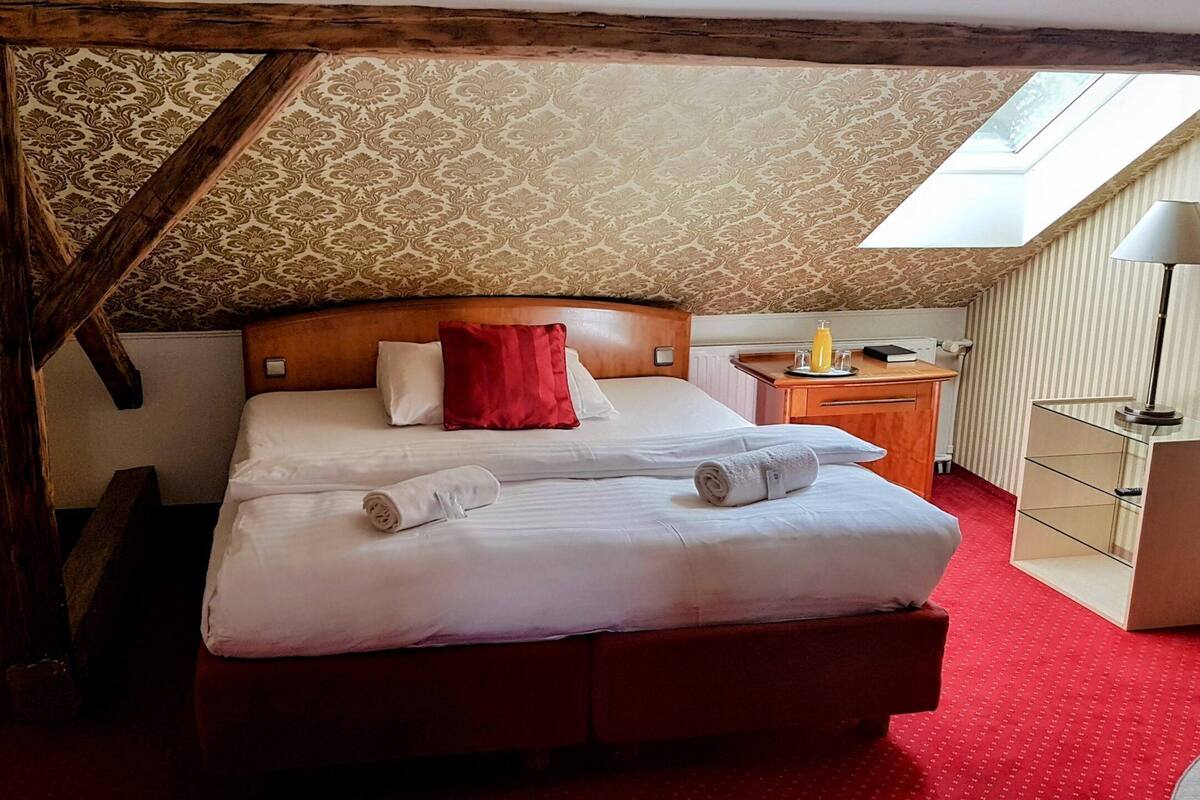 A neatly made bed with white linens and a red pillow, a wooden nightstand with a lamp, and a red carpeted floor.