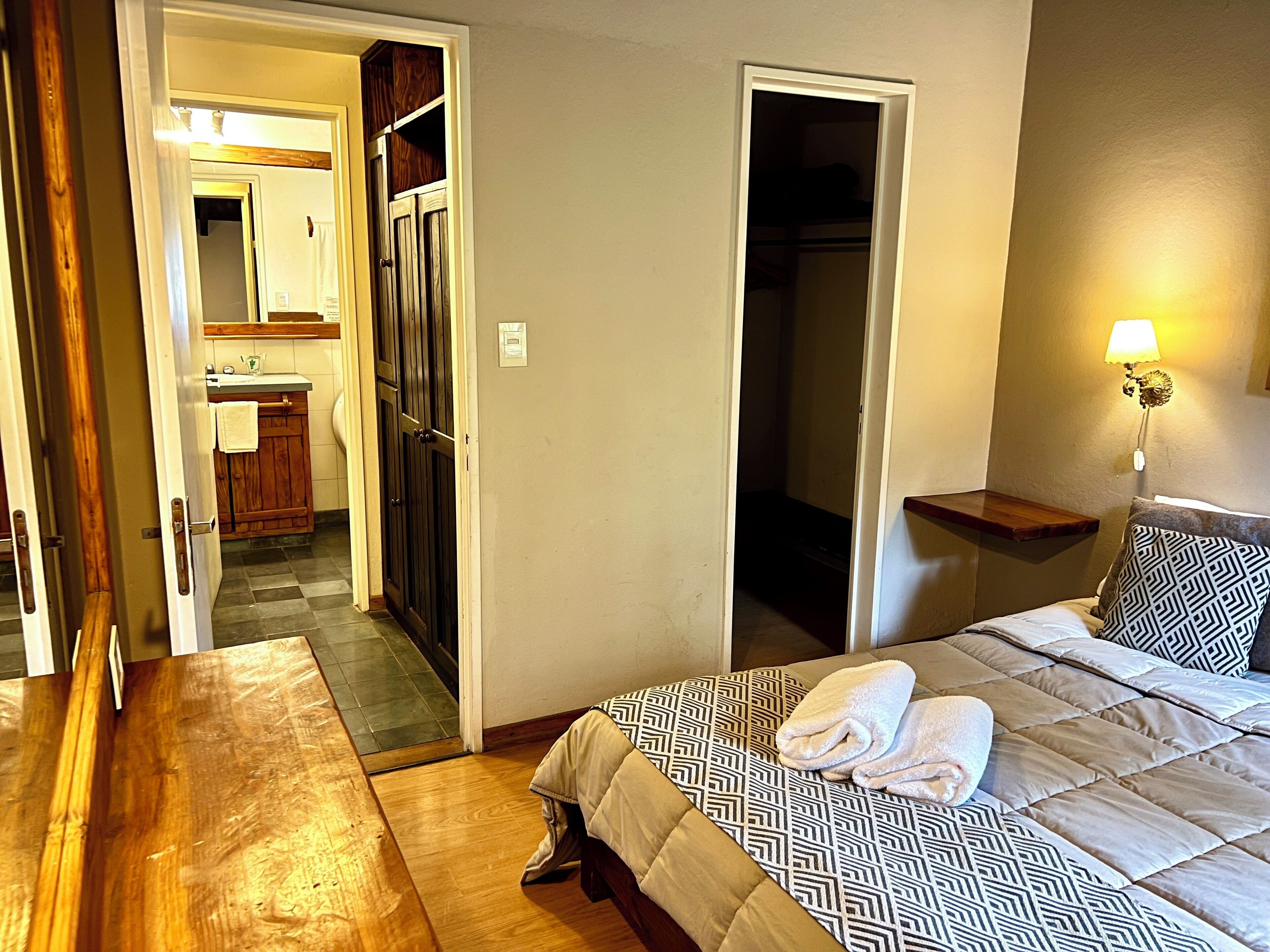 Habitación de hotel con cama, mesita de noche, lámpara y baño visible a través de una puerta abierta.