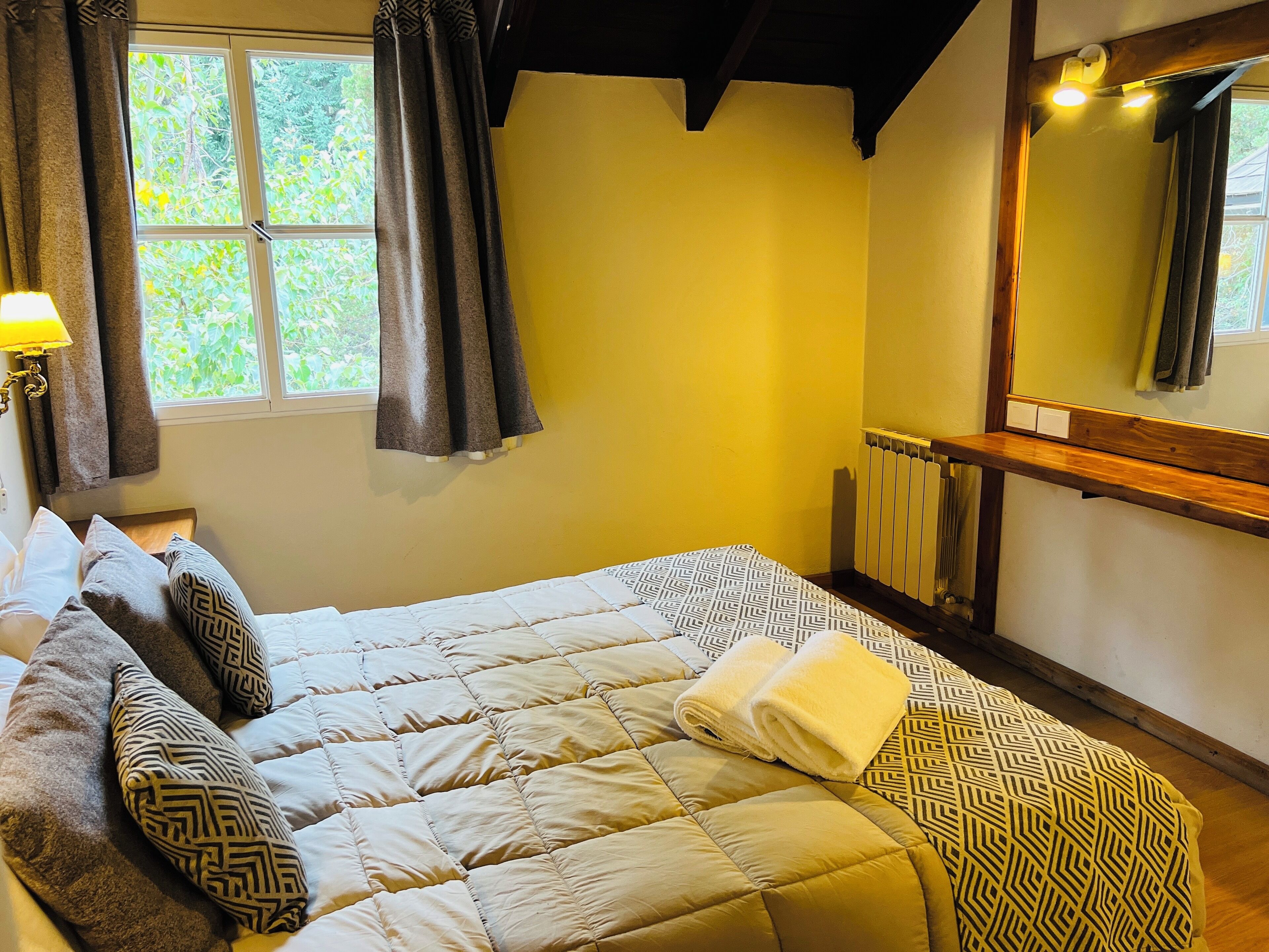 Una cama con una colcha acolchada, almohadas y una toalla enrollada. Hay una ventana con cortinas, un radiador y un espejo en la pared.