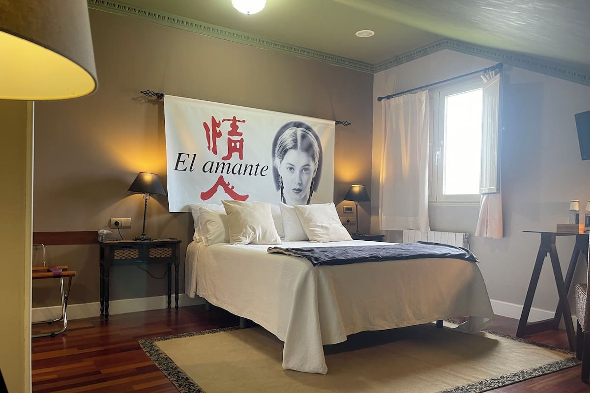 Un dormitorio con una cama, dos mesitas de noche, una lámpara, un cartel, una ventana con cortinas y un radiador.