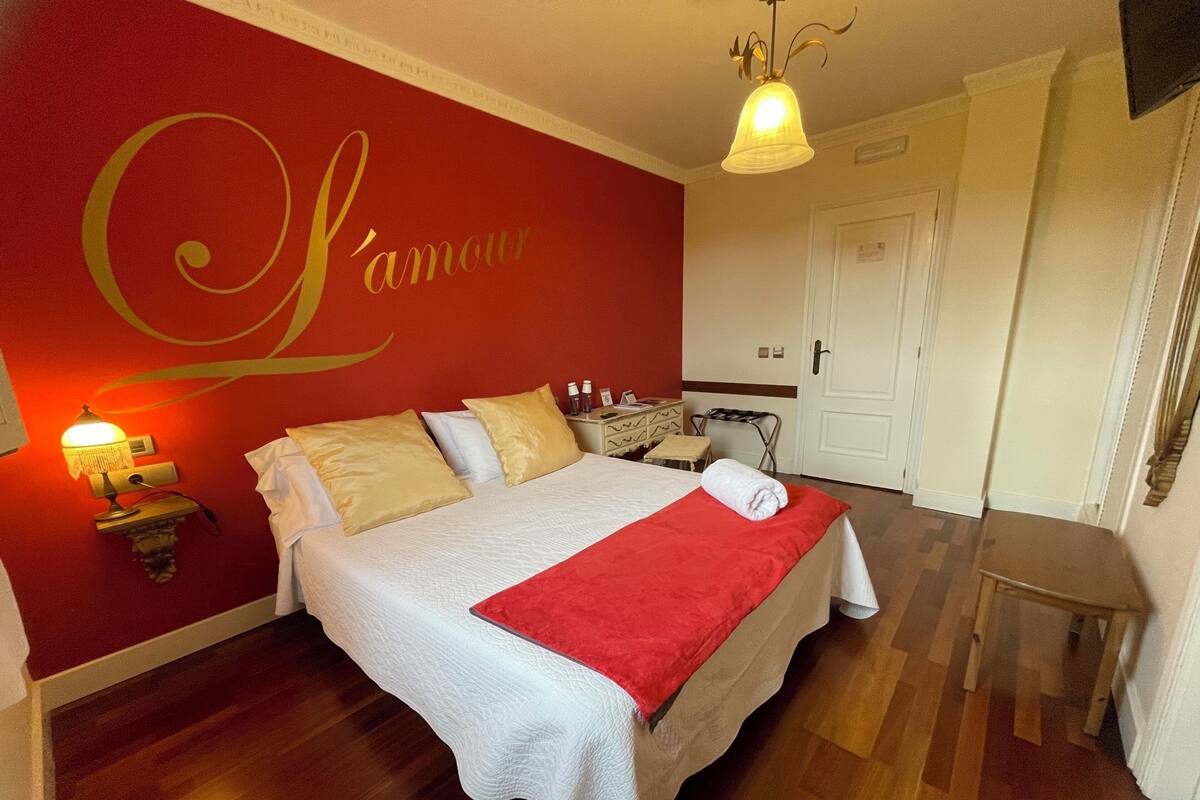 Habitación de hotel con cama, pared roja con la palabra 'L'amour', mesita de noche con lámpara y suelo de madera.