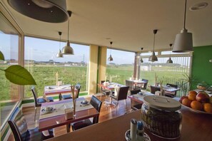 Ein Essbereich mit Tischen und Stühlen, große Fenster mit Blick auf ein grünes Feld und hängende Pendelleuchten.