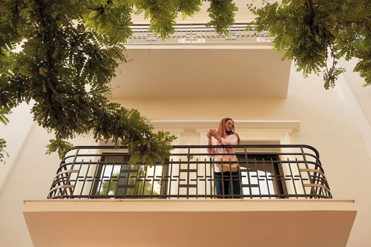 Un balcon avec une rambarde en métal, une personne debout et un arbre aux feuilles vertes.