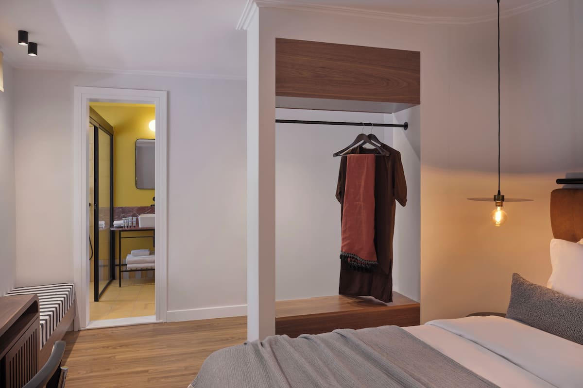 Une chambre moderne avec une tête de lit en bois, un luminaire suspendu et une salle de bain visible, dotée d’un mur jaune et d’un miroir.