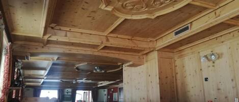 Un intérieur en bois avec des tables dressées pour le repas, un plafond décoratif et une fenêtre laissant entrer la lumière naturelle.