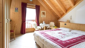 A bedroom with a wooden ceiling, a bed with a patterned bedspread, a chair, a small table with a cup and saucer, and a window with red curtains. - Cevedale (Valfurva)