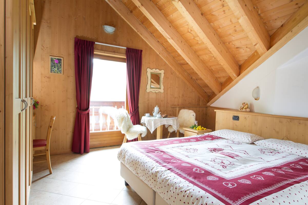 A bedroom with a wooden ceiling, a bed with a patterned bedspread, a chair, a small table with a cup and saucer, and a window with red curtains.