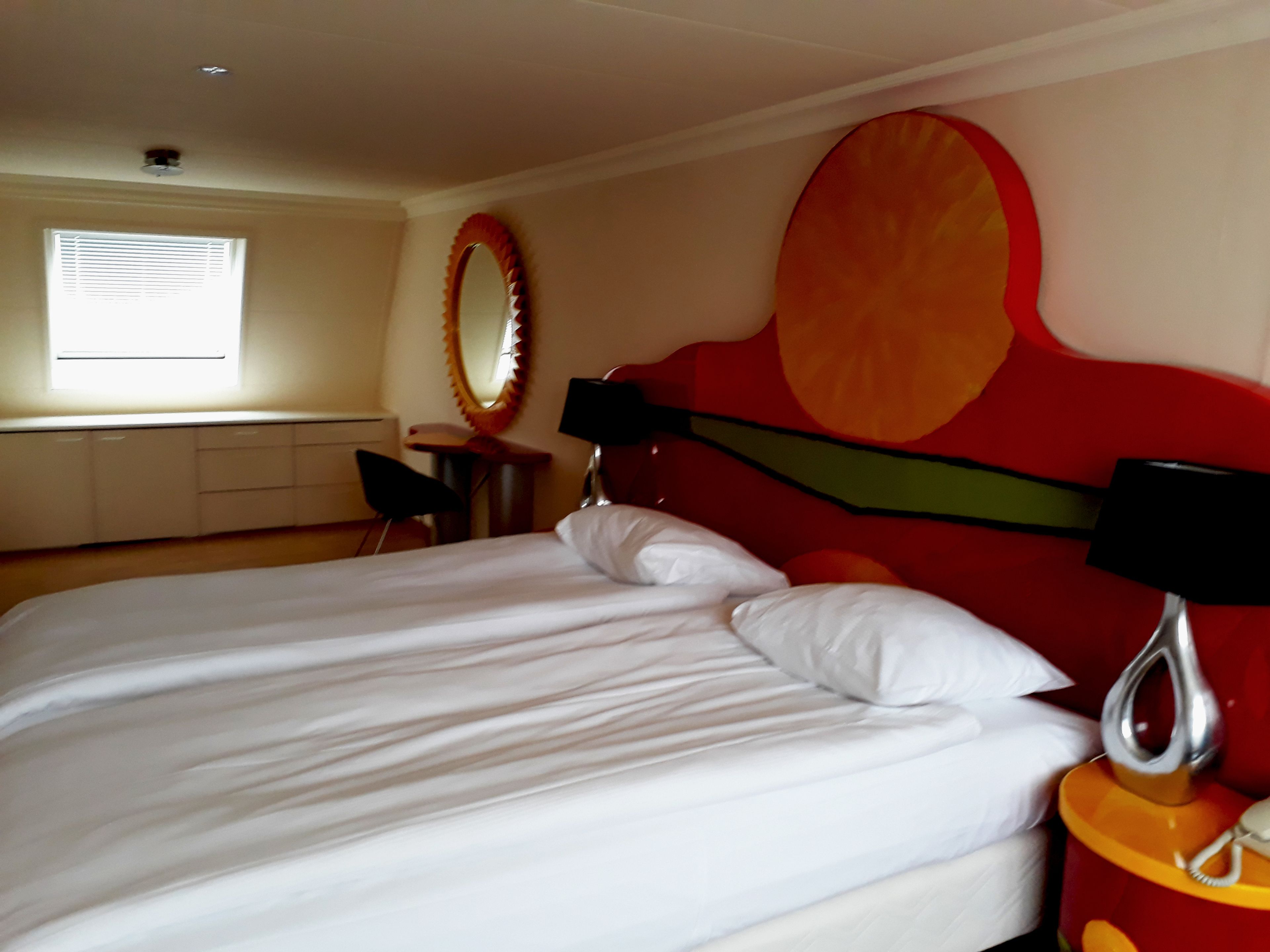 Una habitación de hotel con una cama grande, un espejo redondo, una mesita pequeña y una silla.