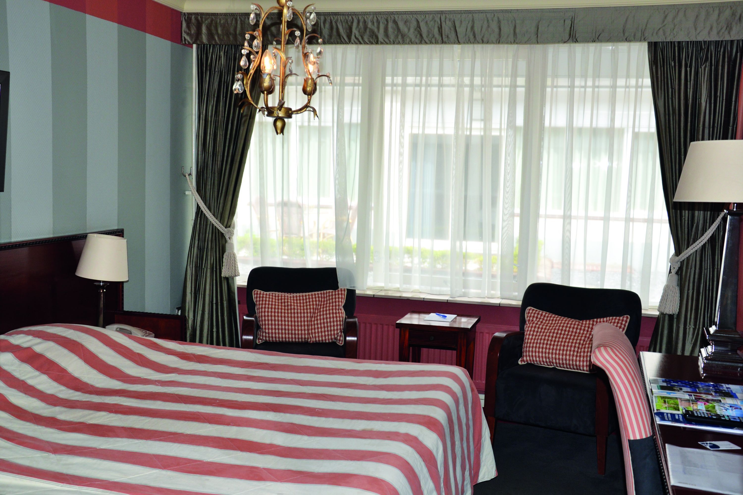 Habitación de hotel con una cama grande, dos sillones, una mesita, una lámpara y un ventanal con cortinas traslúcidas.