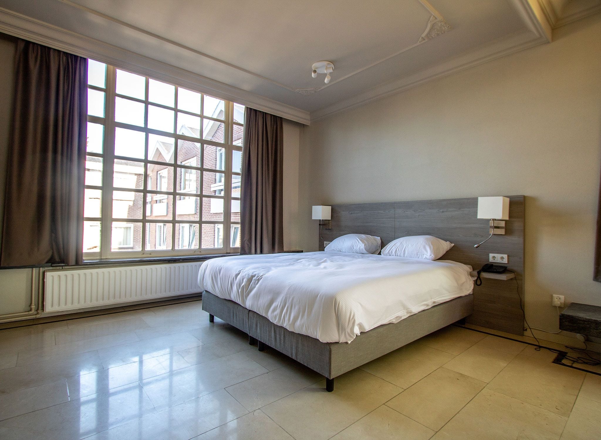 Une chambre d’hôtel avec une grande fenêtre, un lit avec une literie blanche, une table de chevet et une lampe fixée au mur.
