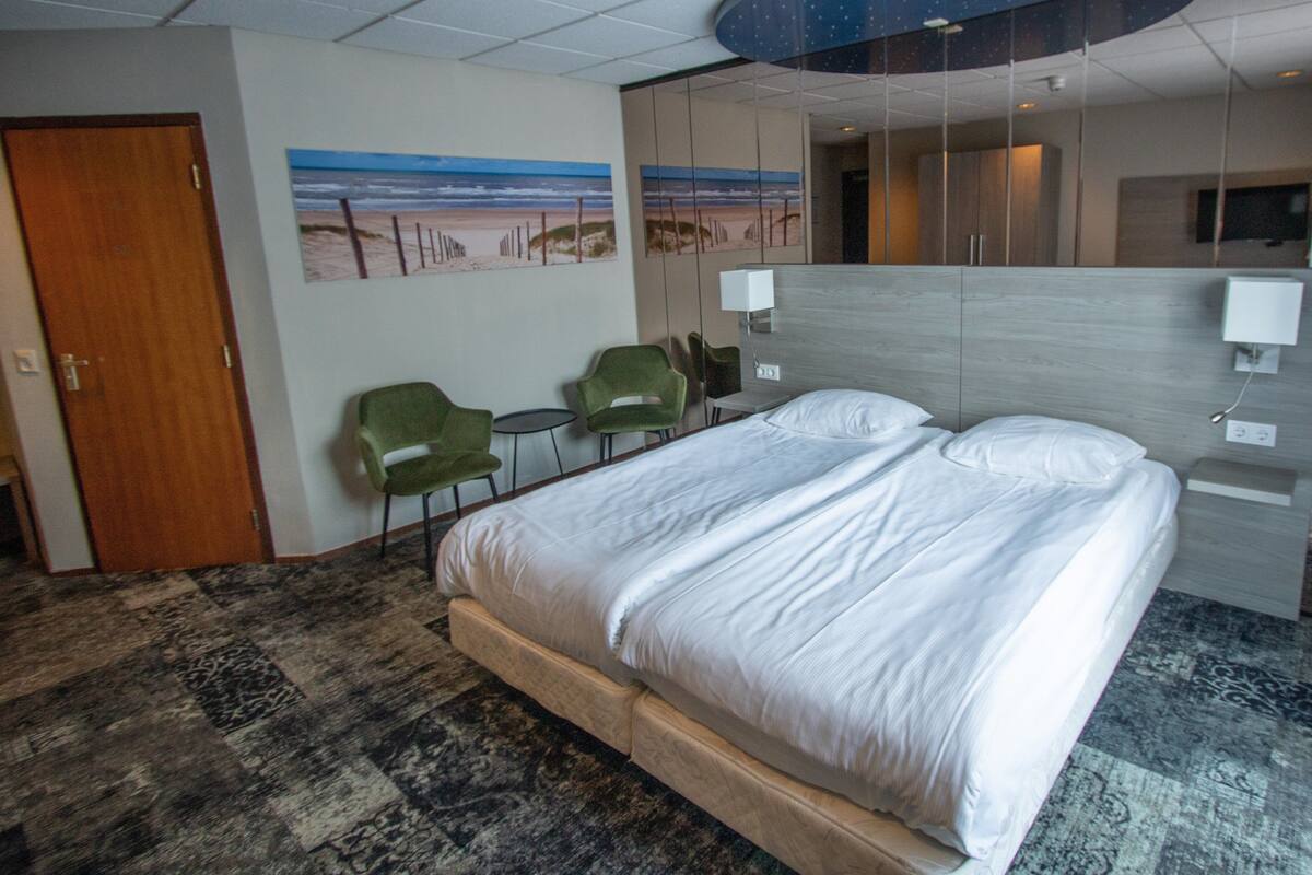 Hotellihuone, jossa on suuri sänky, kaksi vihreää tuolia, pieni pöytä ja seinällä ranta-aiheinen taulu.