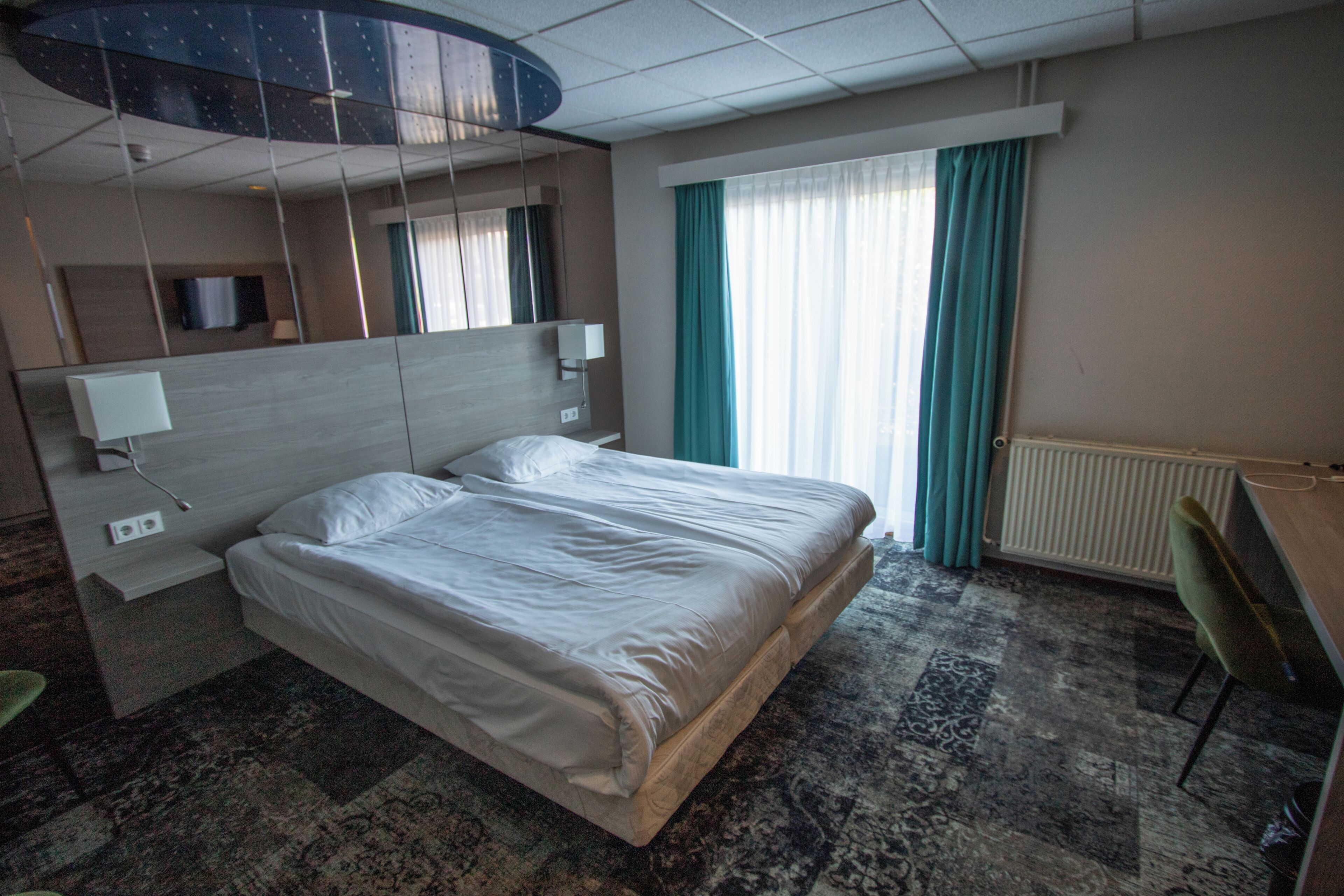 A hotel room with a large bed, a desk, a chair, a mirror, a TV, and a window with curtains.