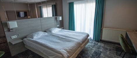 A hotel room with a large bed, a desk, a chair, a mirror, a TV, and a window with curtains.