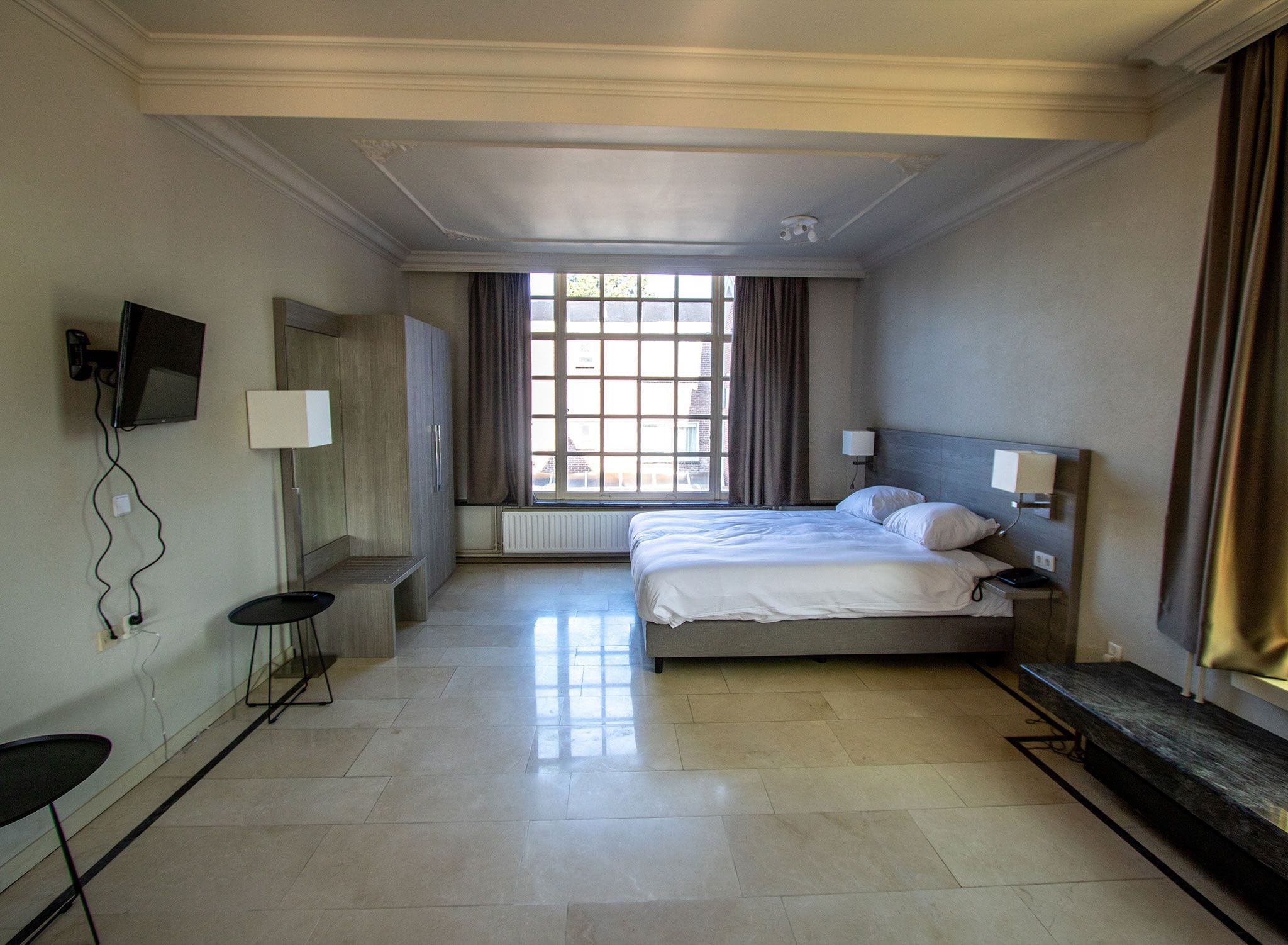 Une chambre d’hôtel avec un lit, des tables de chevet, une télévision, une fenêtre avec des rideaux et un radiateur.