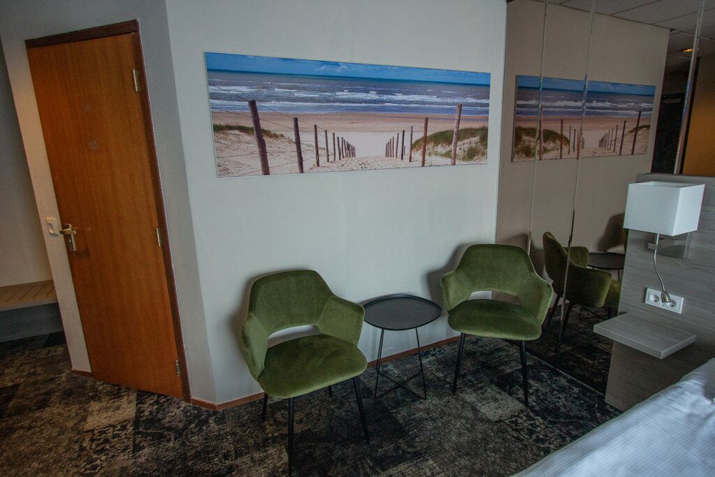 A hotel room with a beach-themed wall art, two green chairs, a small round table, and a bed.