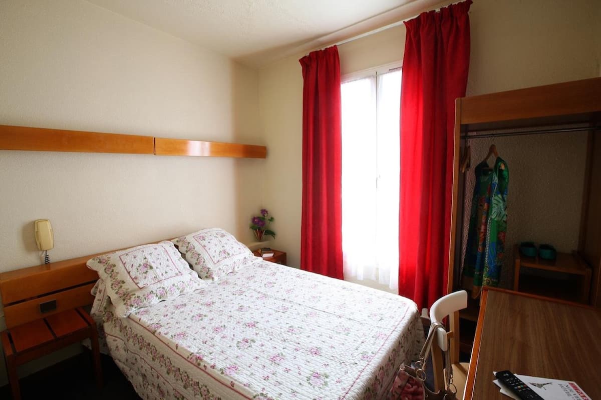 A hotel room with a bed, a bedside table, a nightstand, a chair, a desk, a wardrobe, and a window with red curtains.
