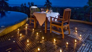 An outdoor dining area with a table and chairs under a tree, surrounded by candles and a view of the ocean. - Bali Nibbana Resort (Seririt)