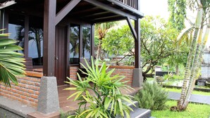 A covered porch with a brick wall and wooden beams, surrounded by lush greenery and a small pond. - Bali Nibbana Resort (Seririt)