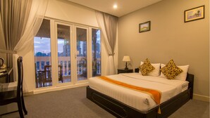 A hotel room with a large bed, a chair, a nightstand, a lamp, and a view of the city through a sliding door. - Diamond Palace Hotel (Phnom Penh)