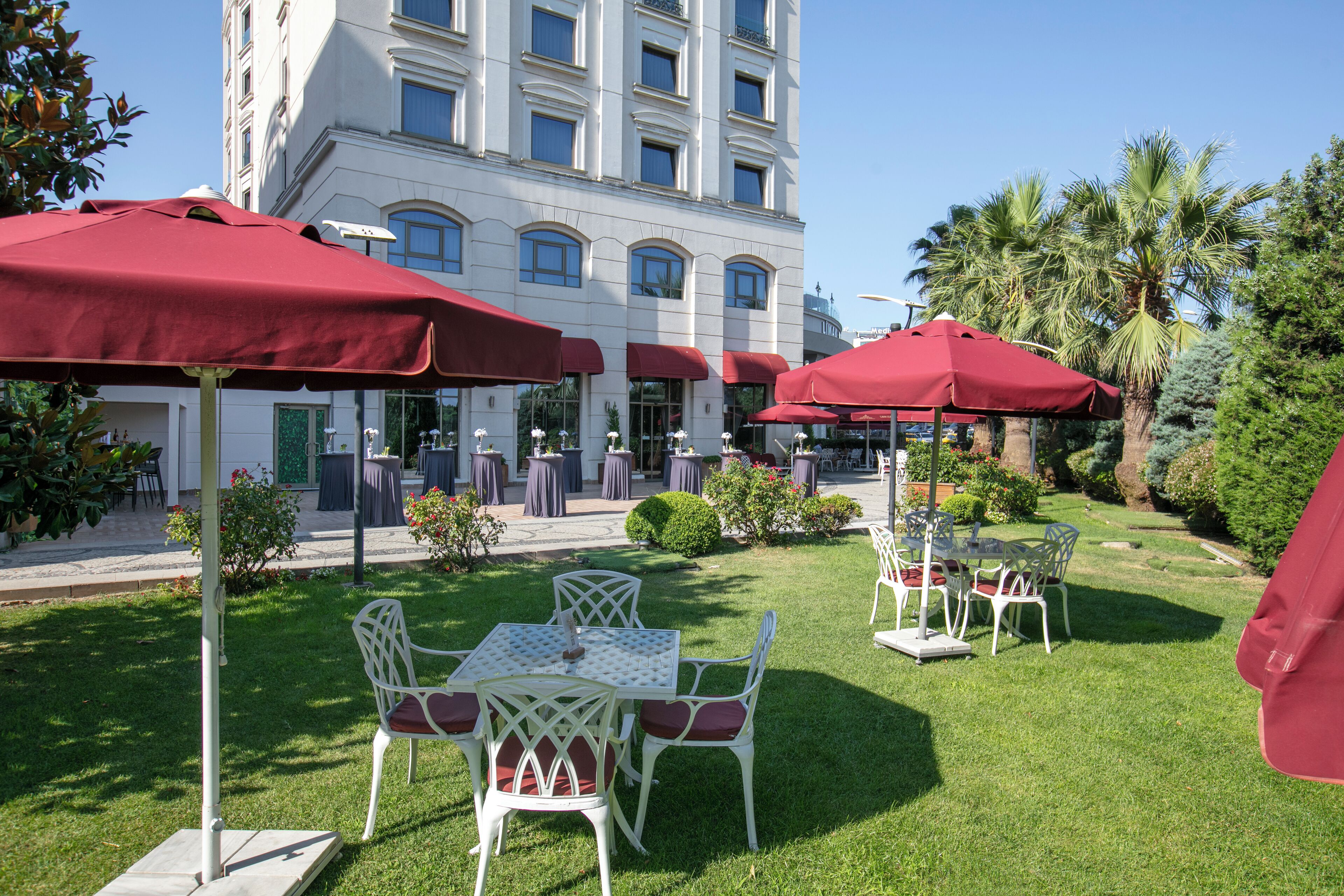 Un espace extérieur aménagé avec des chaises blanches et des parasols rouges, entouré d’une pelouse bien entretenue et de palmiers.