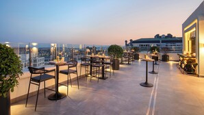 Une terrasse sur le toit, avec des espaces de détente en plein air, des plantes en pot et une vue sur la ville au crépuscule.