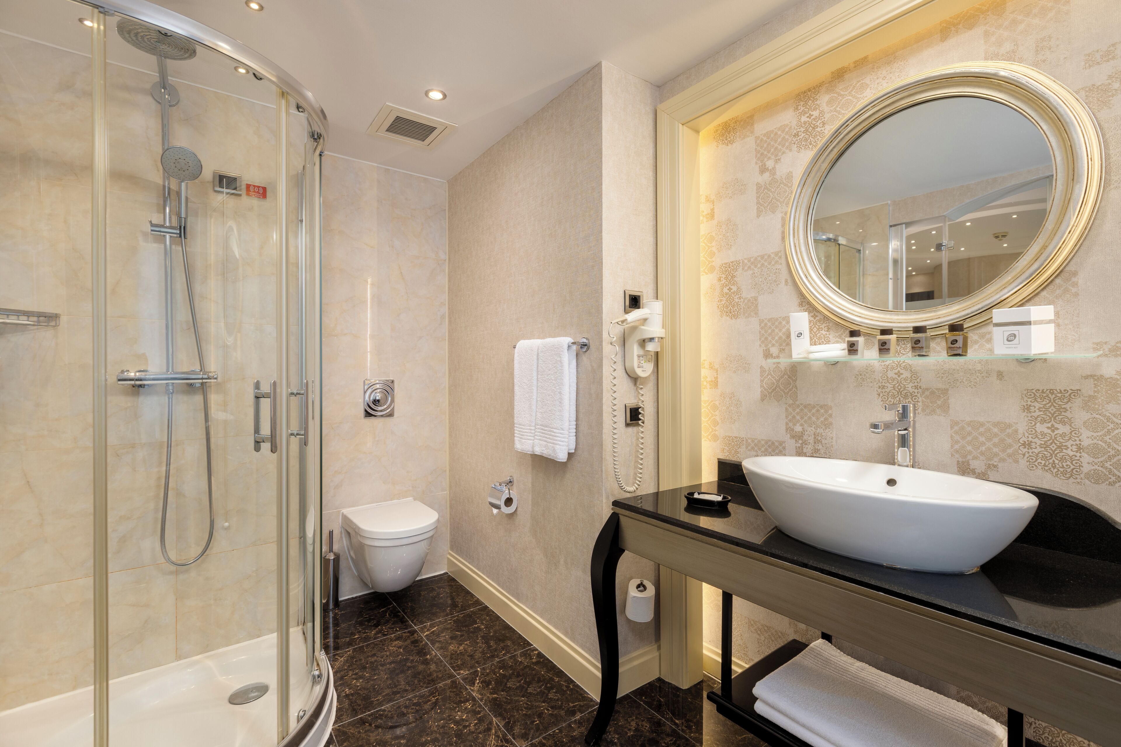 A modern bathroom with a glass-enclosed shower, a white toilet, a vanity with a large oval mirror, and a black countertop.