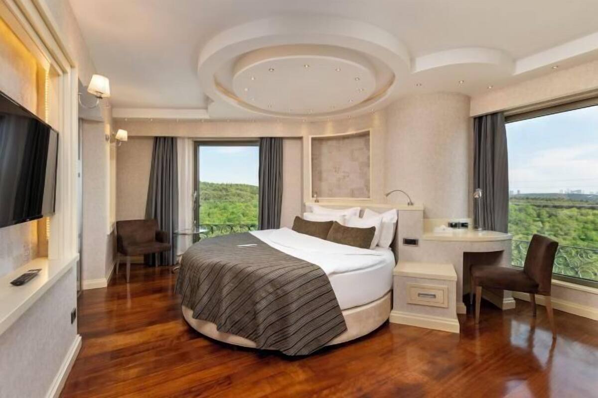Een slaapkamer met een groot bed, een bureau, een stoel, een televisie en uitzicht op het groen.