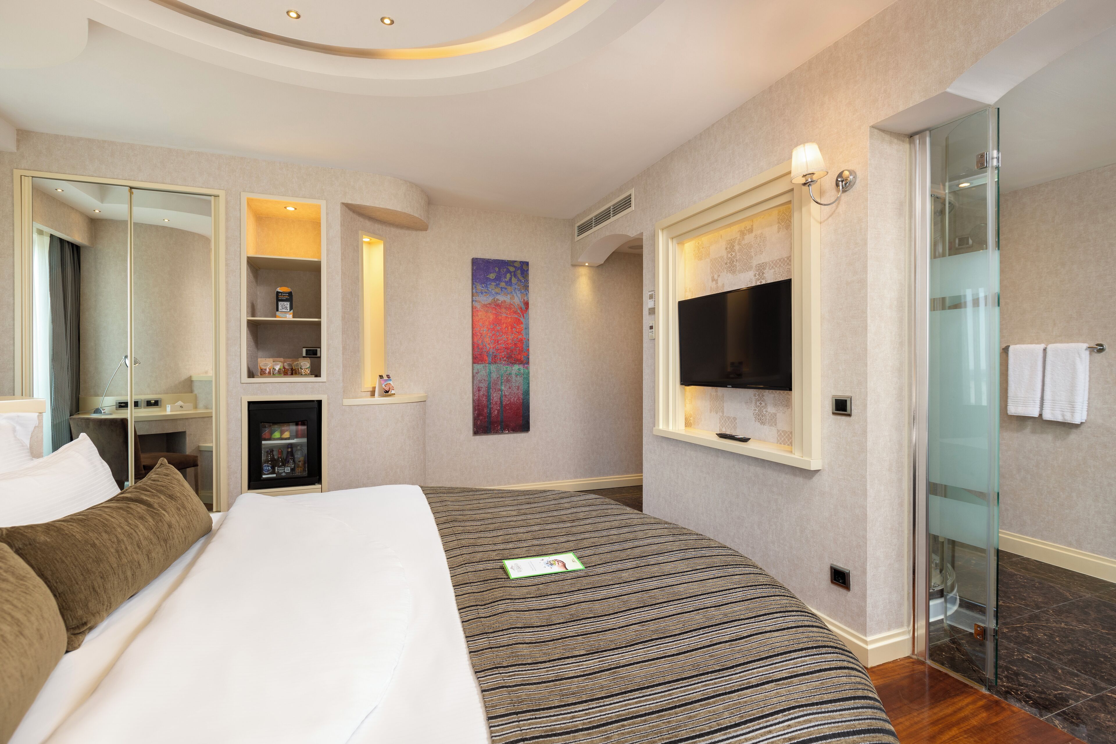 Une chambre d’hôtel dotée d’un grand lit, d’une télévision, d’une douche en verre et d’un petit coin salon avec un bureau et des étagères.