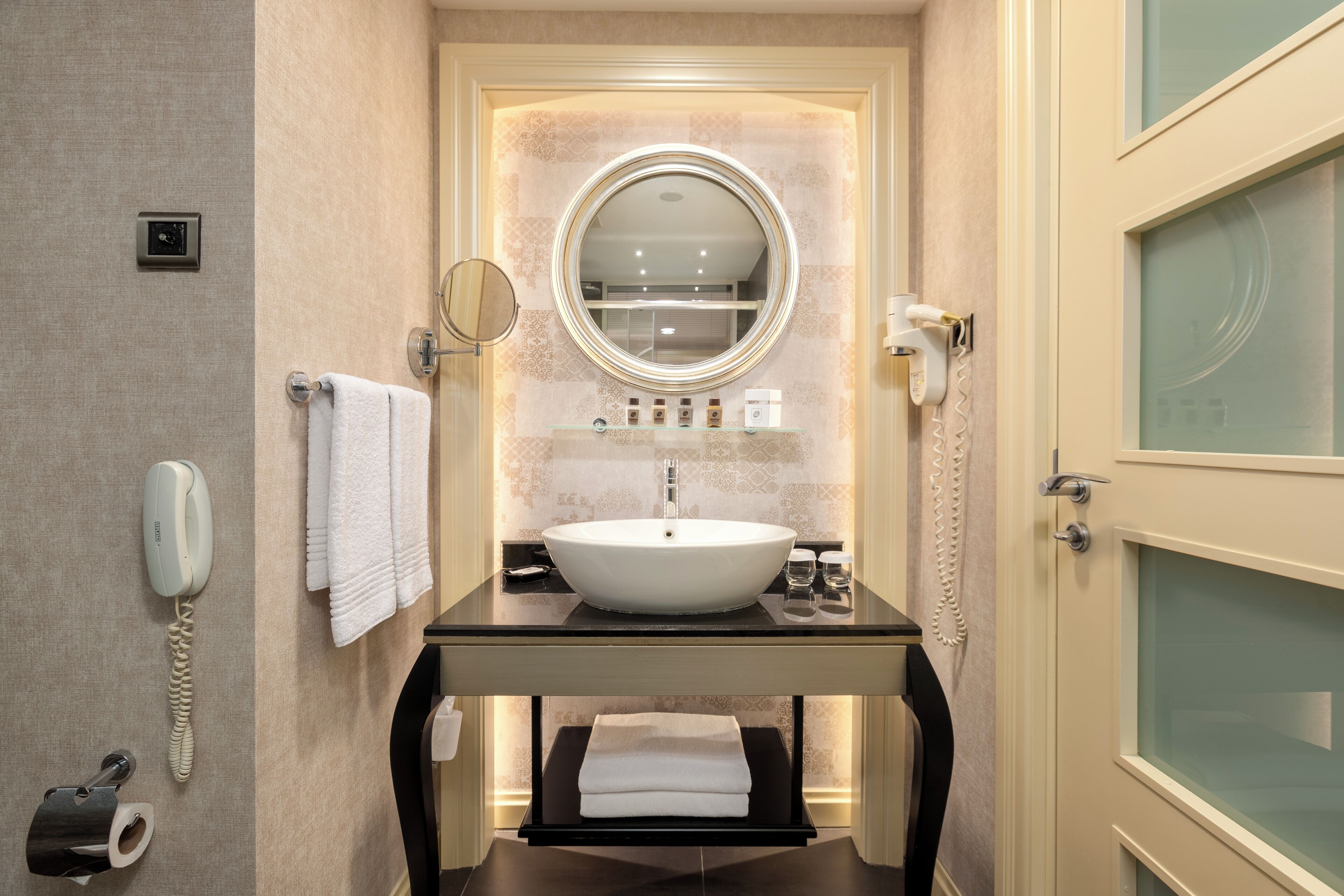 A bathroom with a round mirror, a white sink, a towel rack, a telephone, and a glass-enclosed door.