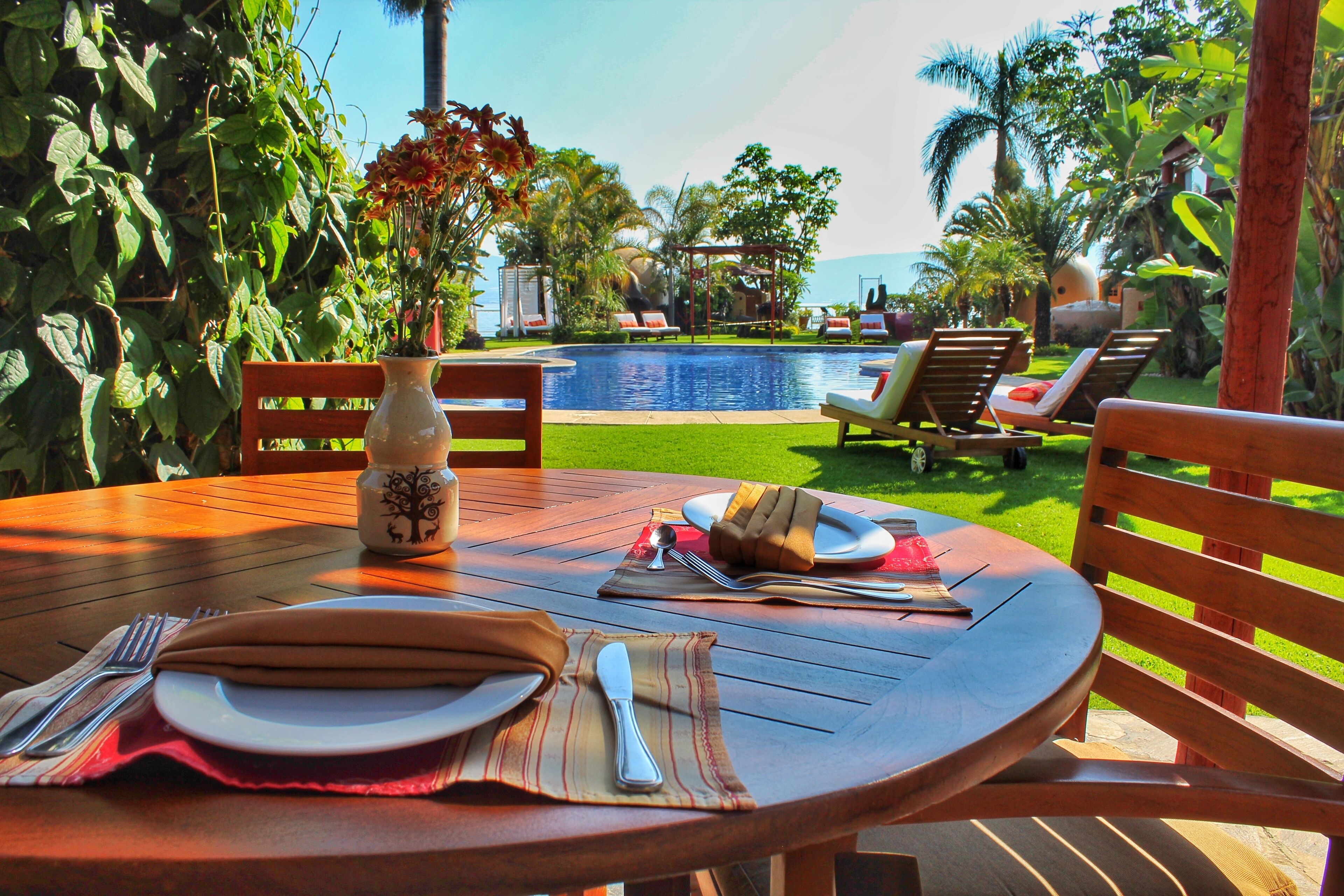 Mesa de jantar posta ao ar livre, com vista para uma piscina e palmeiras.