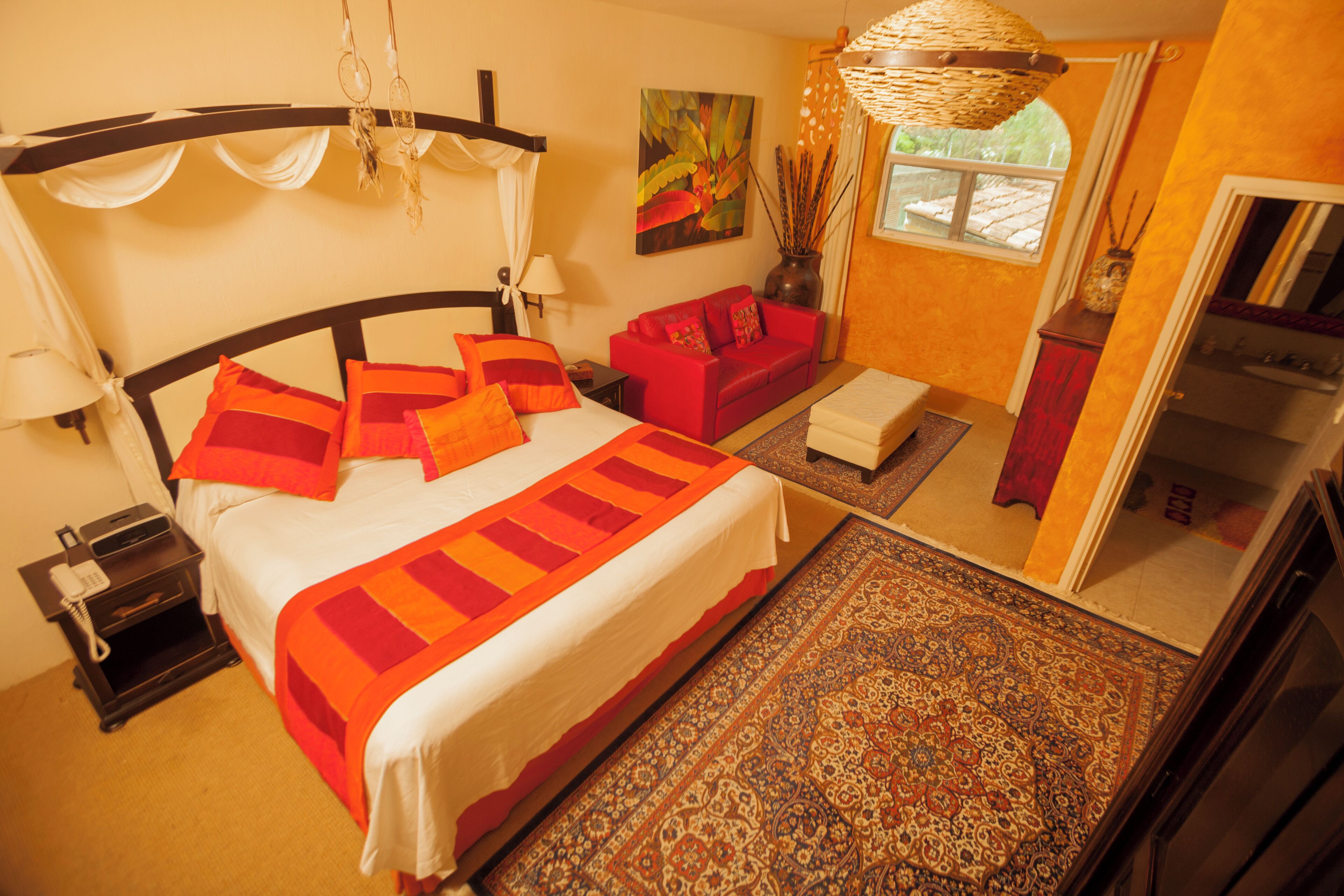 A bedroom with a bed, a red sofa, a nightstand, a lamp, a painting, a window, and a hanging light fixture.