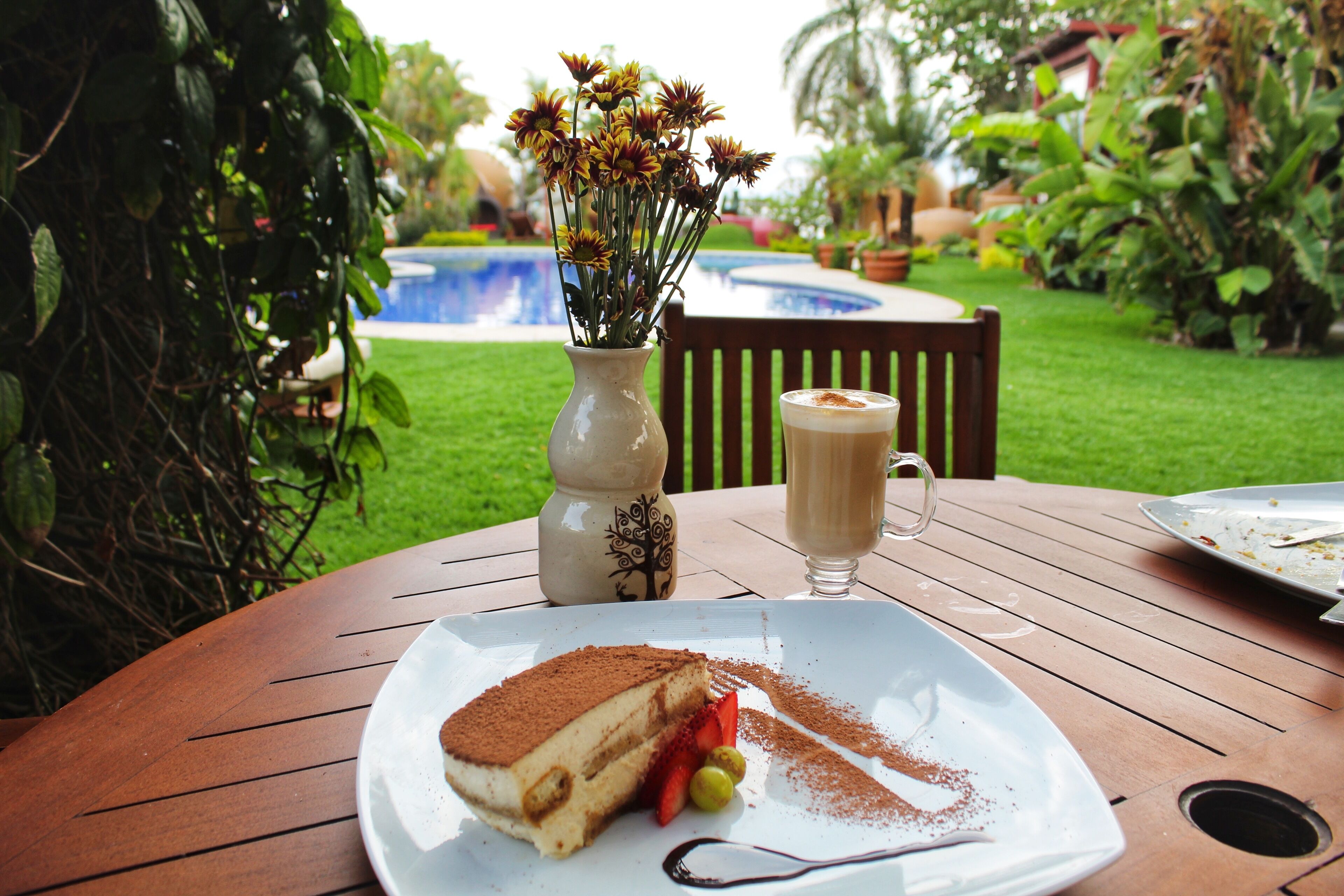Una mesa de comedor con un plato de tiramisú, una taza de café y un jarrón de flores.
