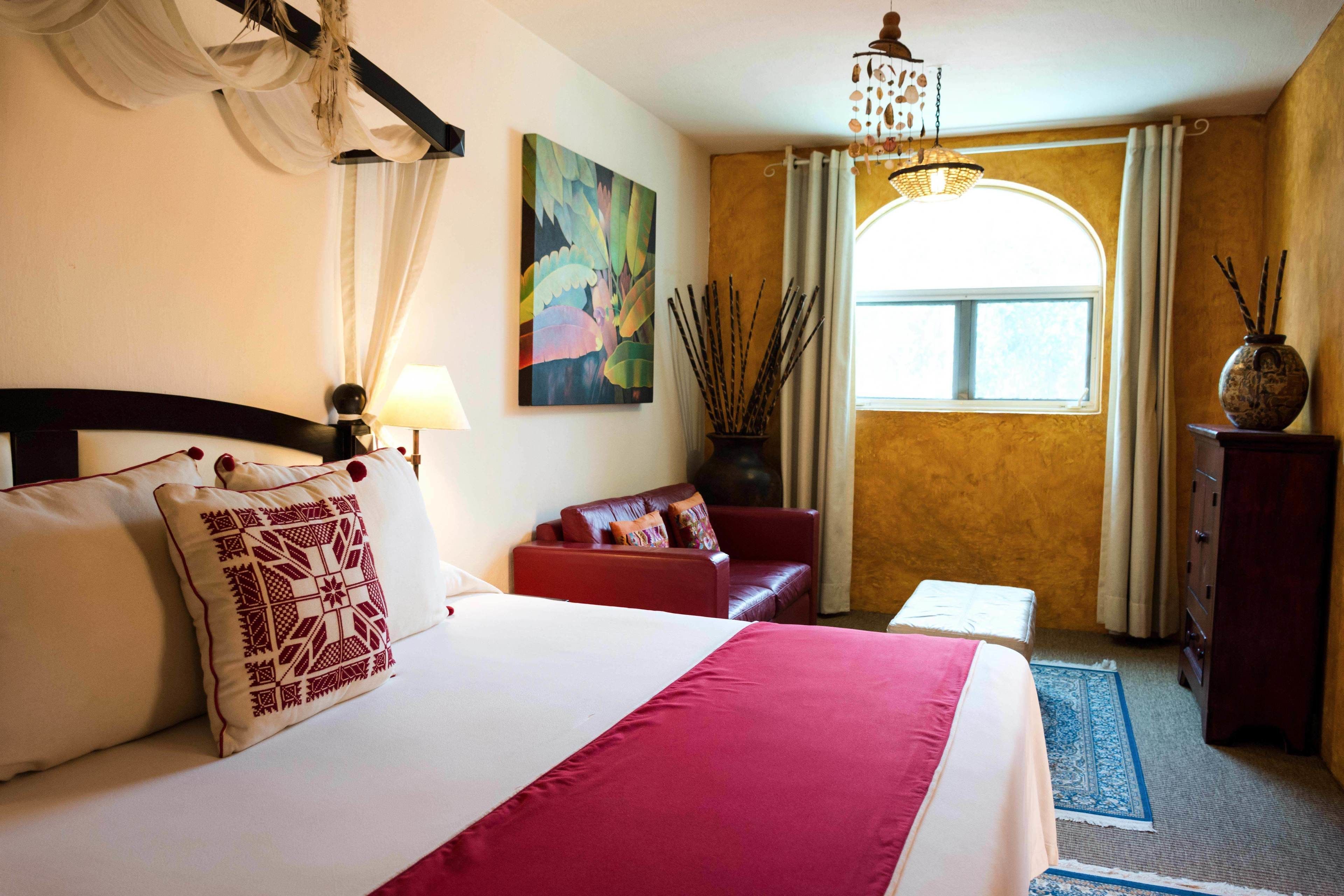 A hotel room with a large bed, a red sofa, a small table, a vase with sticks, and a window with curtains.