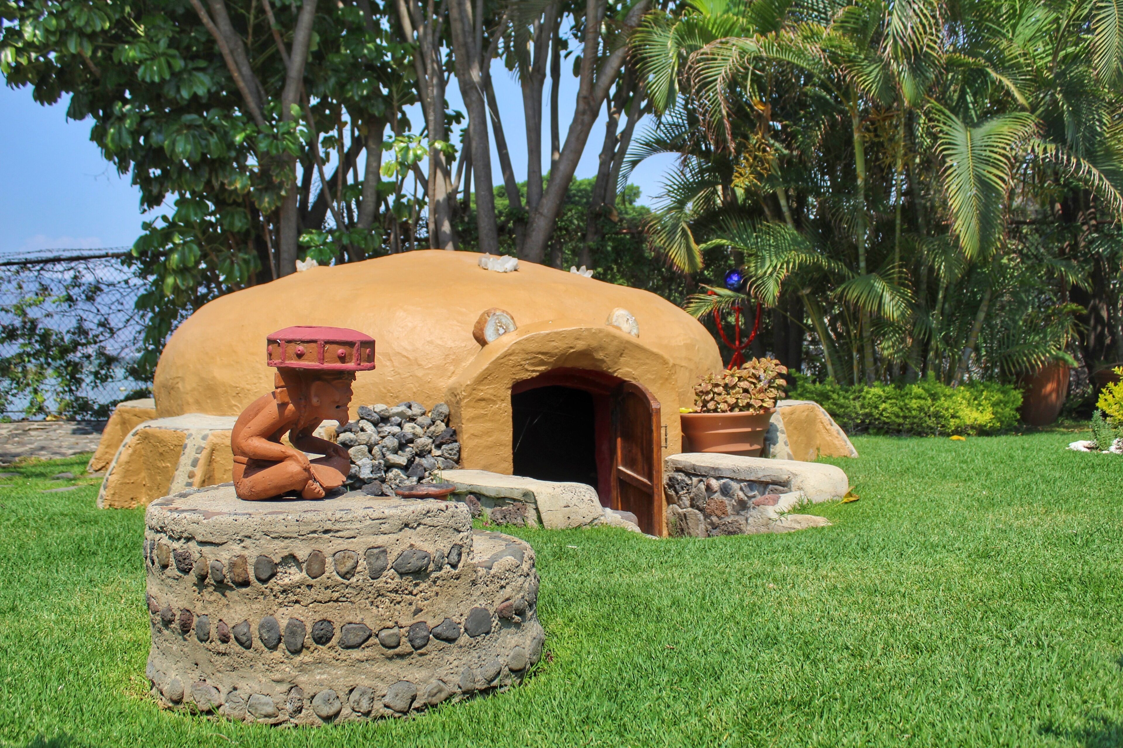 A traditional clay house with a thatched roof, surrounded by a well-maintained lawn and palm trees.