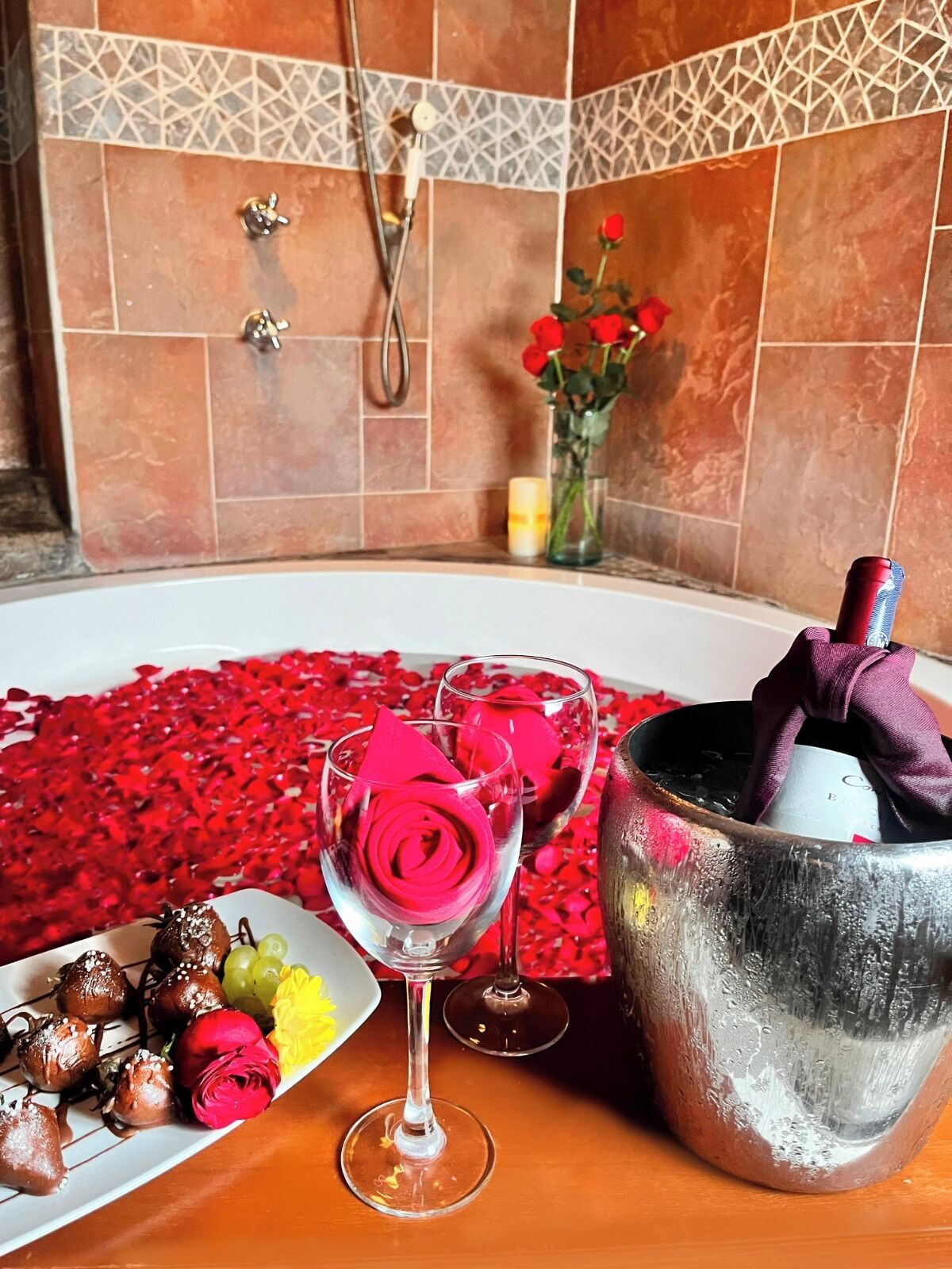 A bathtub filled with rose petals, a glass with a rose, a champagne bottle, and a plate with chocolates and grapes.