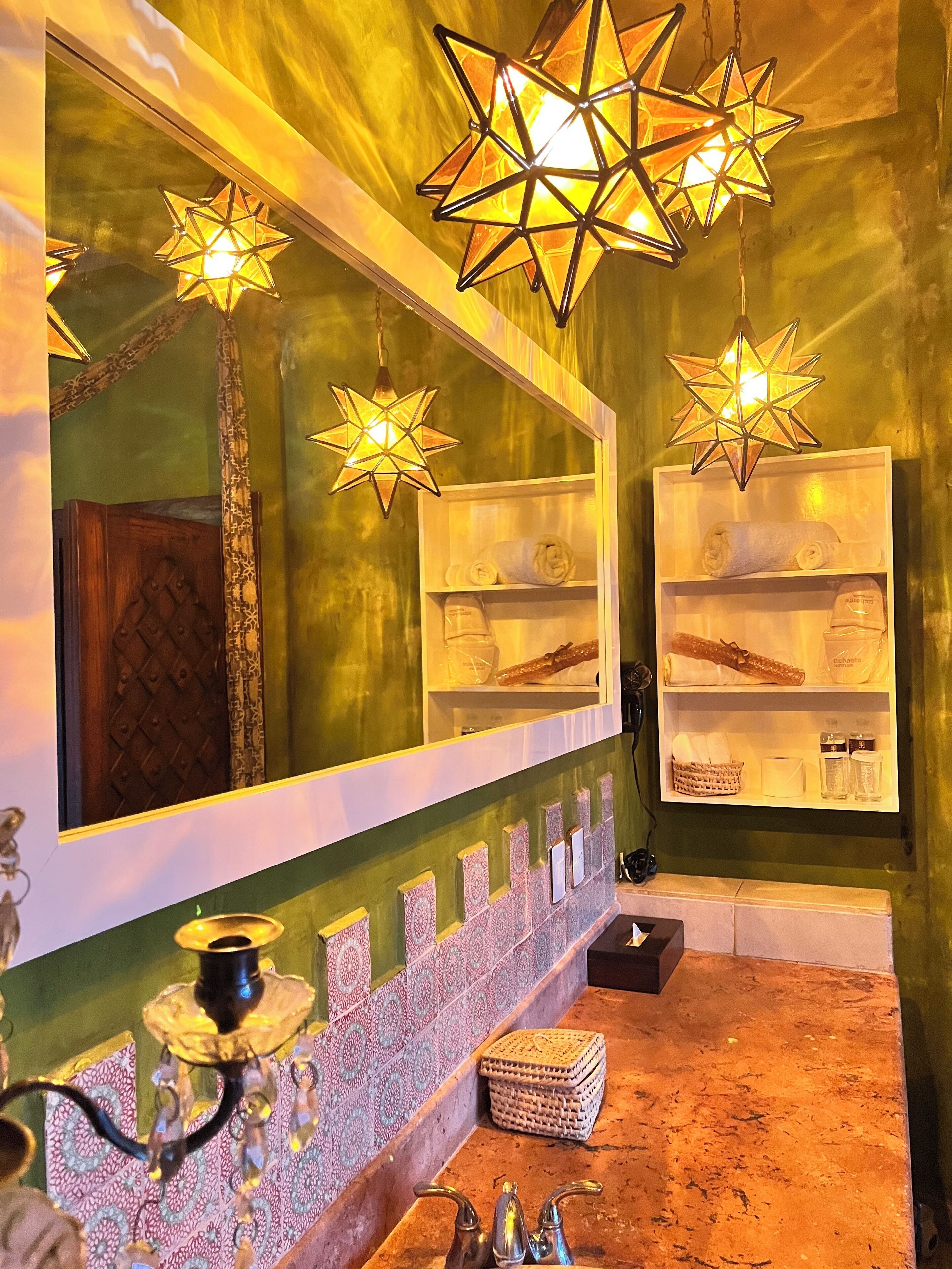 Un baño con un espejo grande, lámparas colgantes con forma de estrella, un salpicadero de azulejos estampados y un tocador de madera.