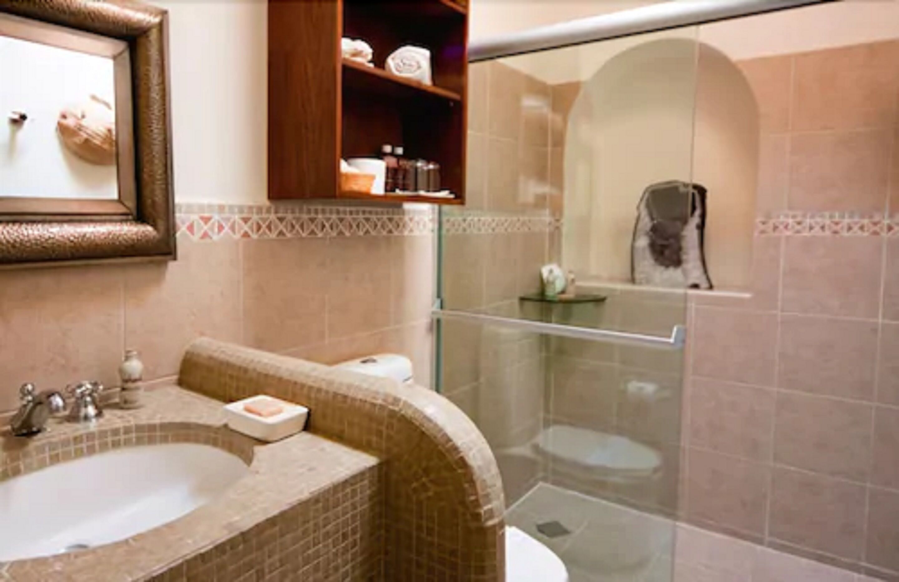 A bathroom with a sink, toilet, and shower. There is a framed picture on the wall and a shelf with toiletries.