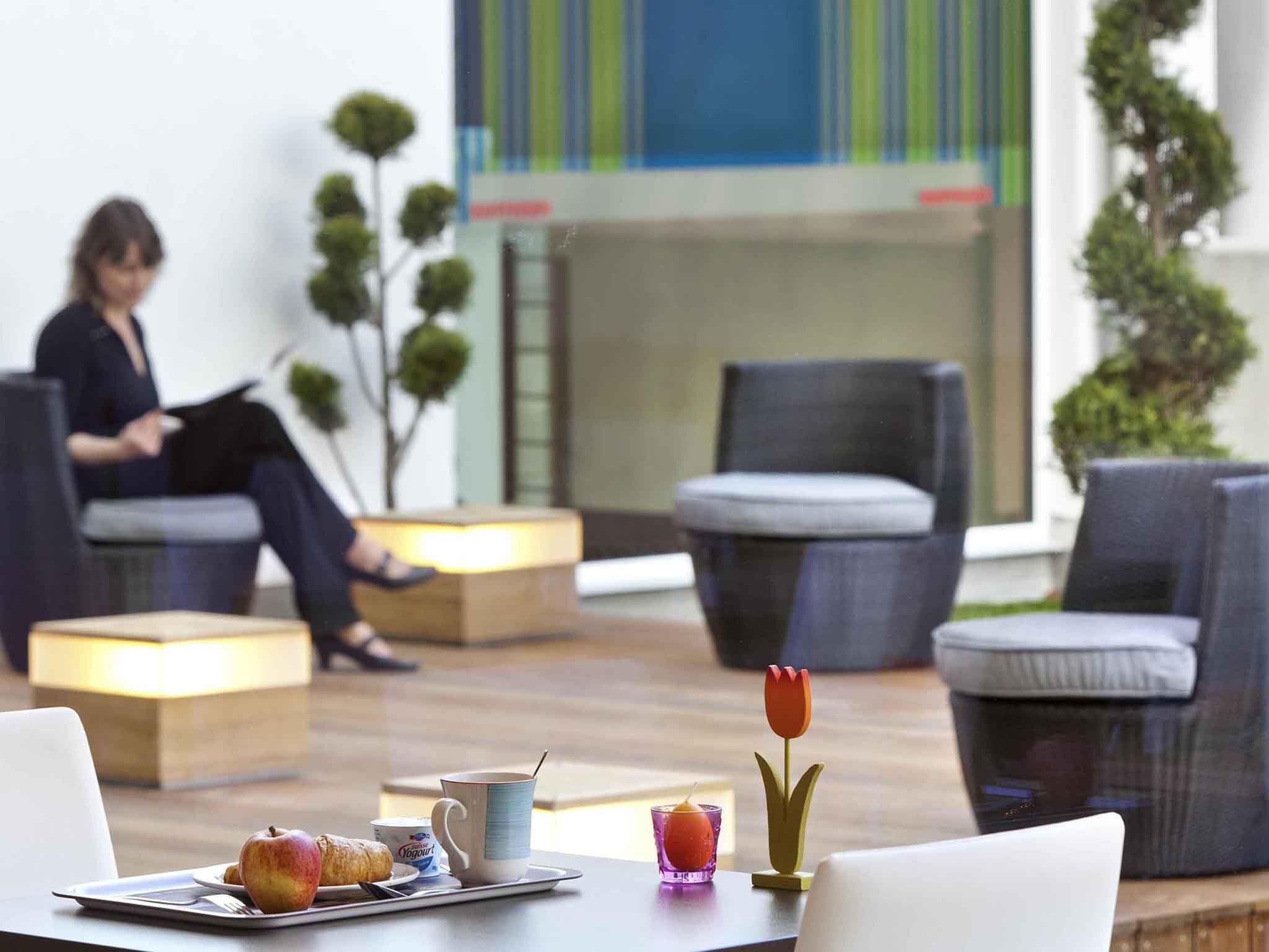 A modern hotel lobby with seating areas, a coffee table with breakfast items, and a person in the background.